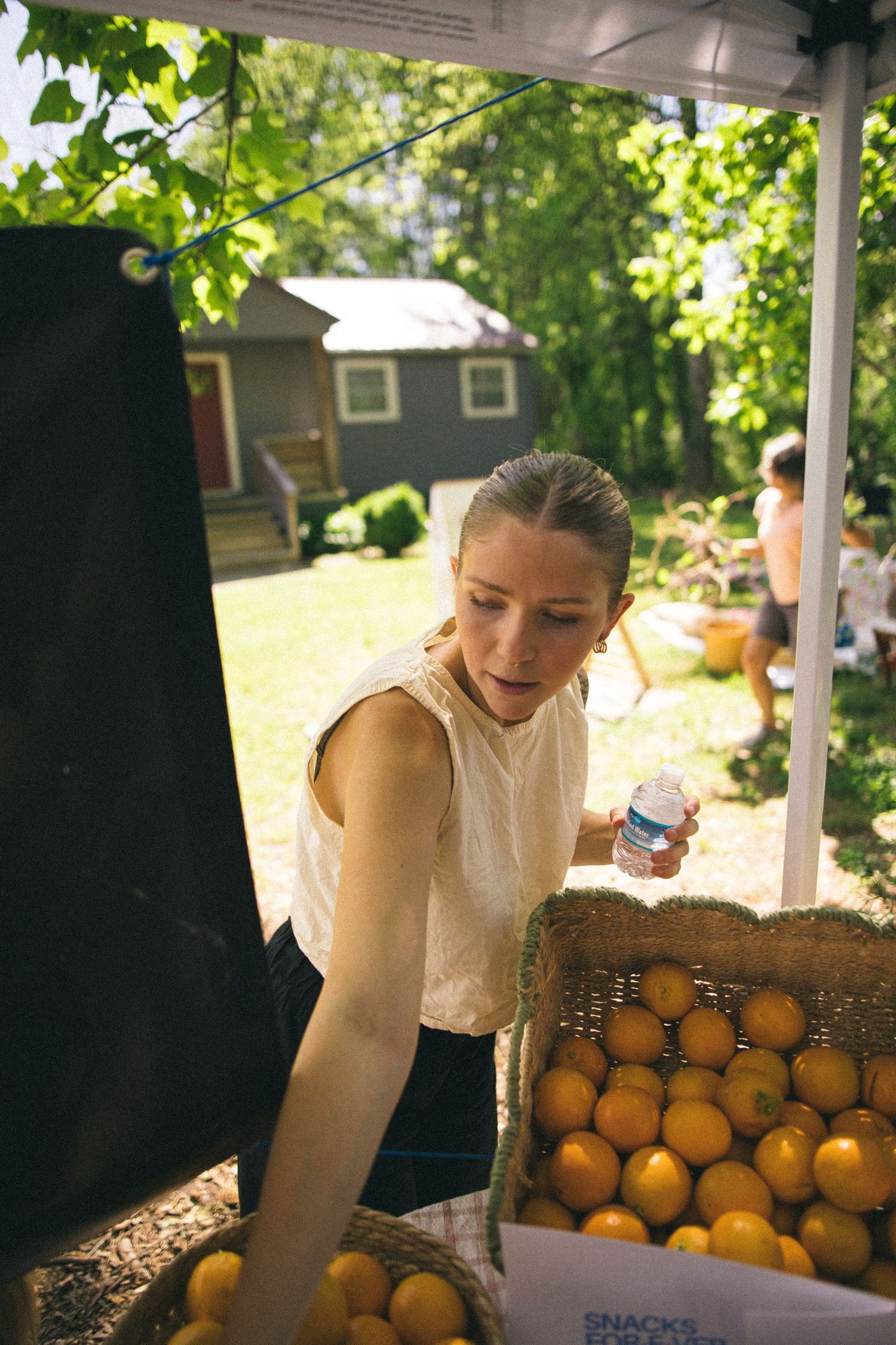 Woman selecting fresh oranges from a rustic burlap basket at an outdoor farmers market on a sunny day.