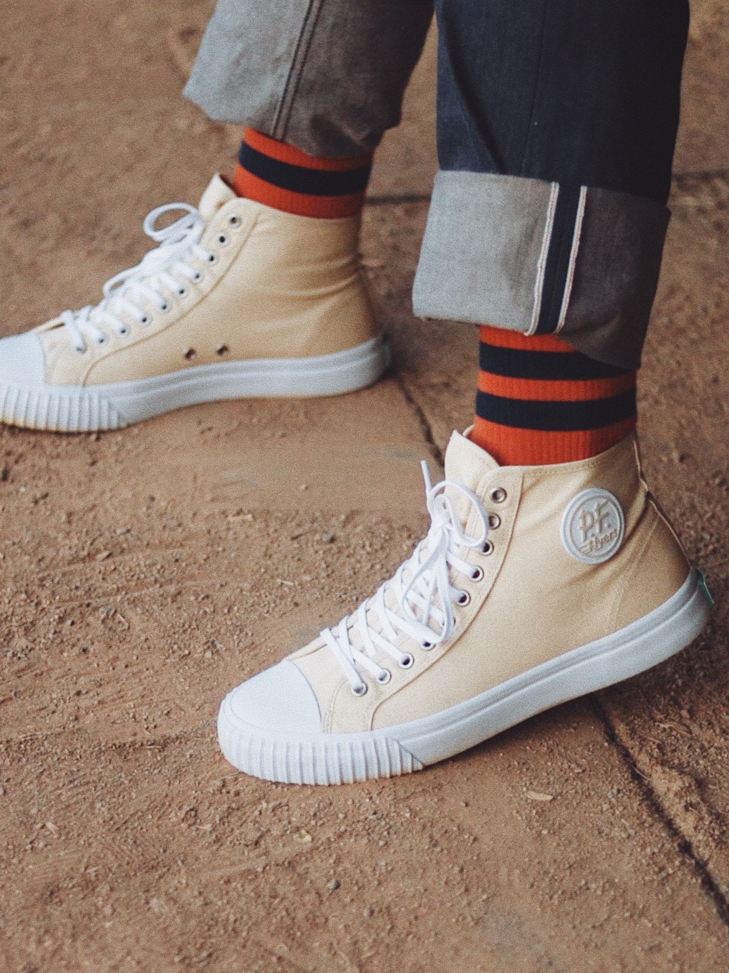 Person wearing cream high-top PF Flyers sneakers with red and black striped socks and cuffed gray jeans.