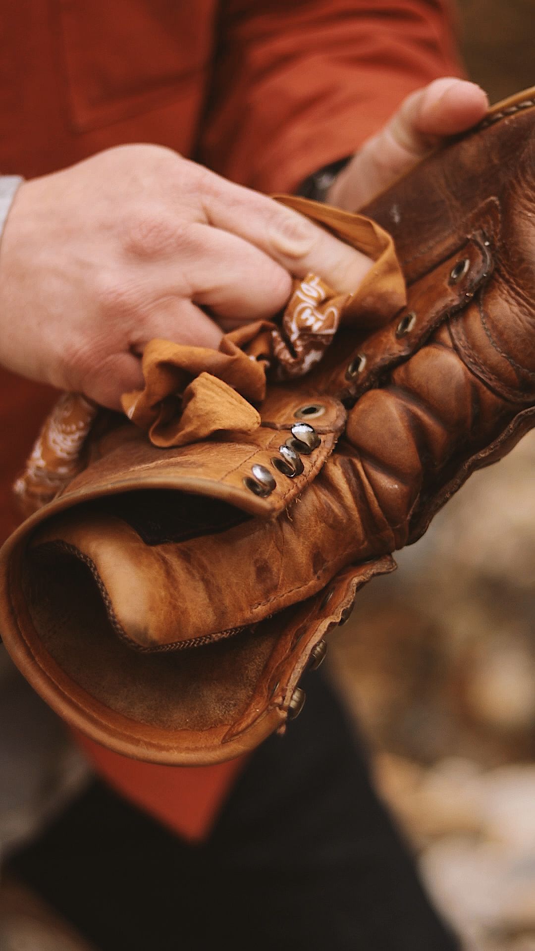 Hands polishing a worn brown leather boot with a cloth, wearing a rust-colored jacket in an outdoor setting.