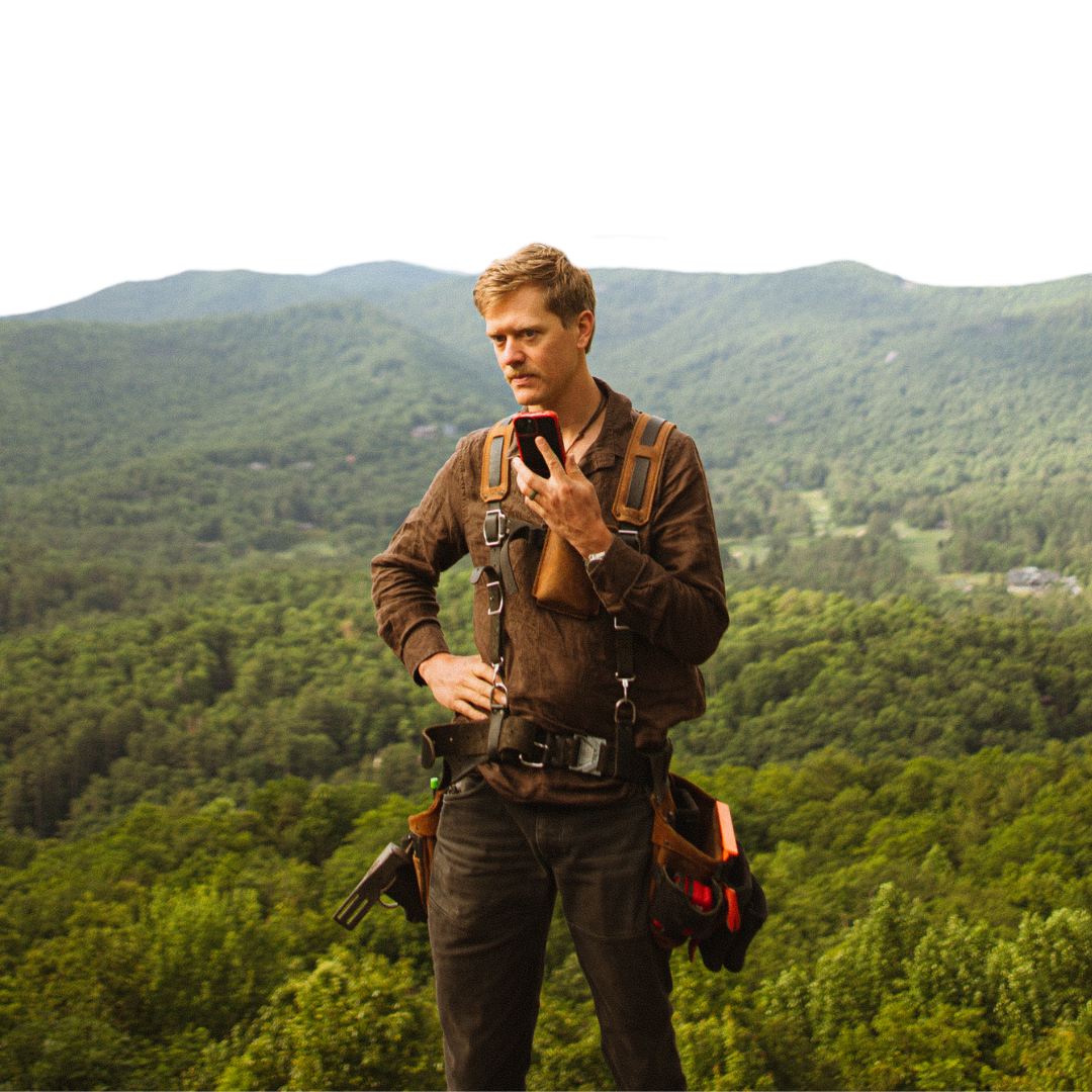 Man in a brown jacket with gear straps checks his phone while standing on a mountain overlook above a forested valley.