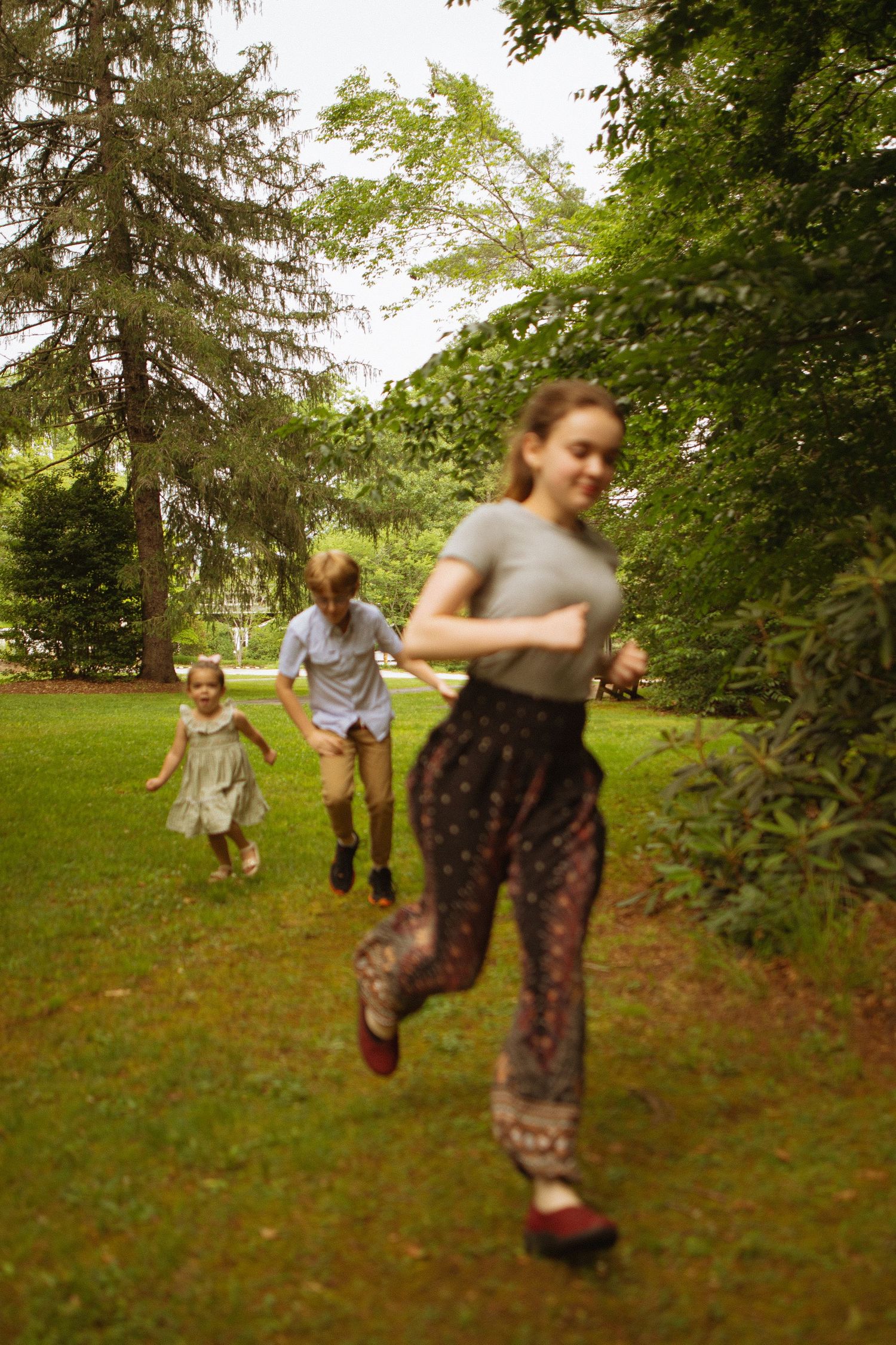 Three children running and playing in a lush green park surrounded by tall trees on a cloudy day.