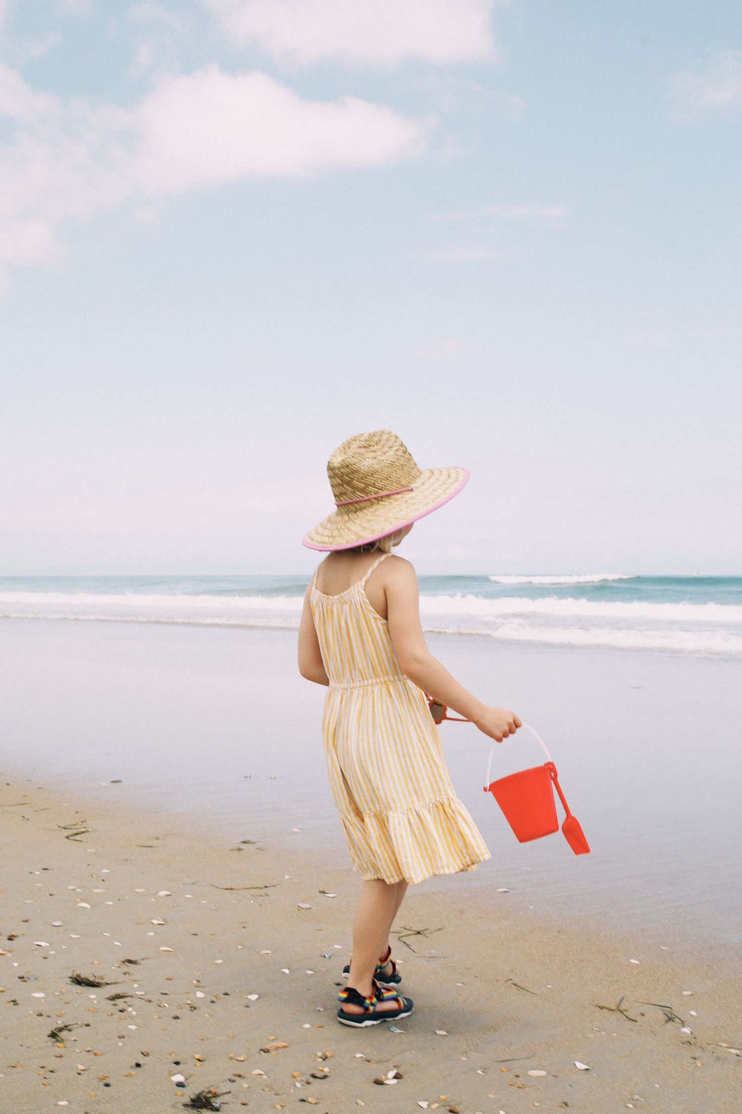 Young girl in yellow dress and straw hat carrying red bucket on sandy beach with ocean waves.