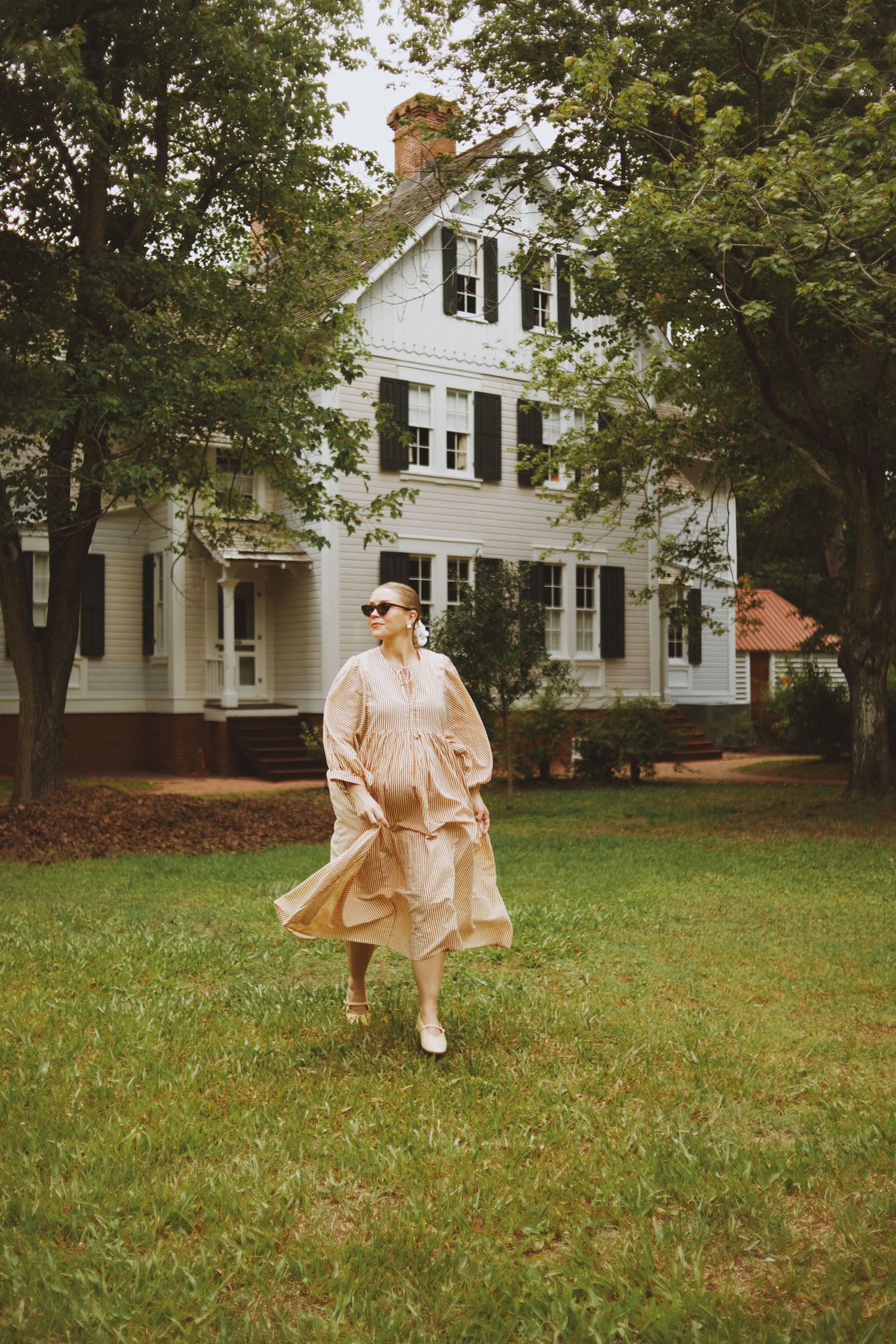 Woman in beige ruffled dress and sunglasses walking on green lawn in front of white colonial house.