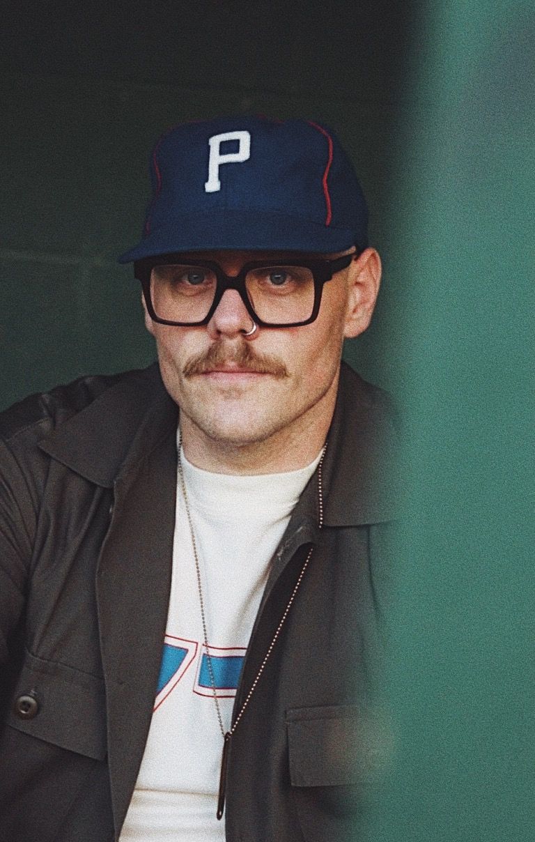 Man with mustache wearing black square glasses, nose ring, blue P cap, and dark jacket poses for portrait photo.