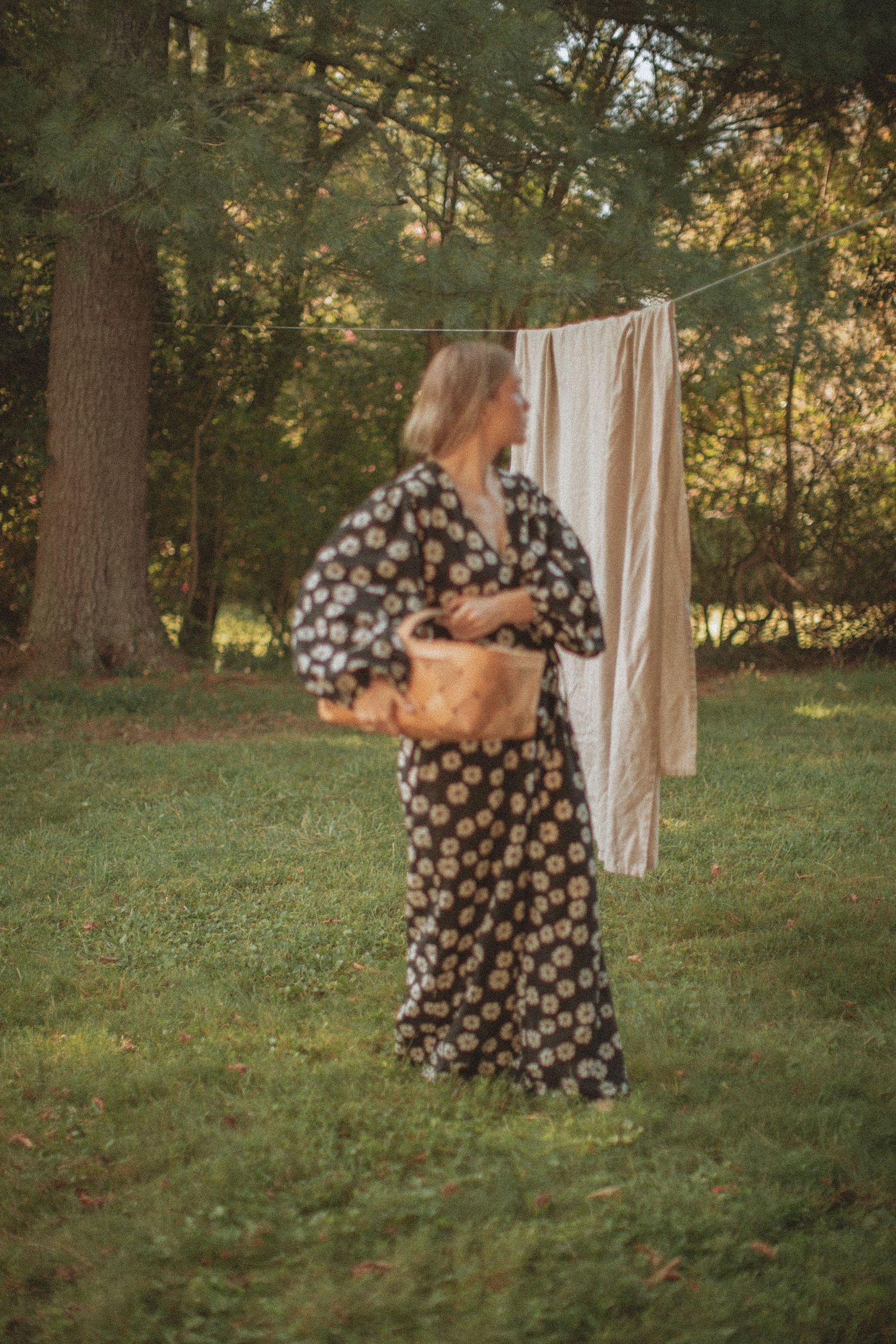 Woman in floral maxi dress holding a wicker basket standing near a clothesline in a lush green backyard.