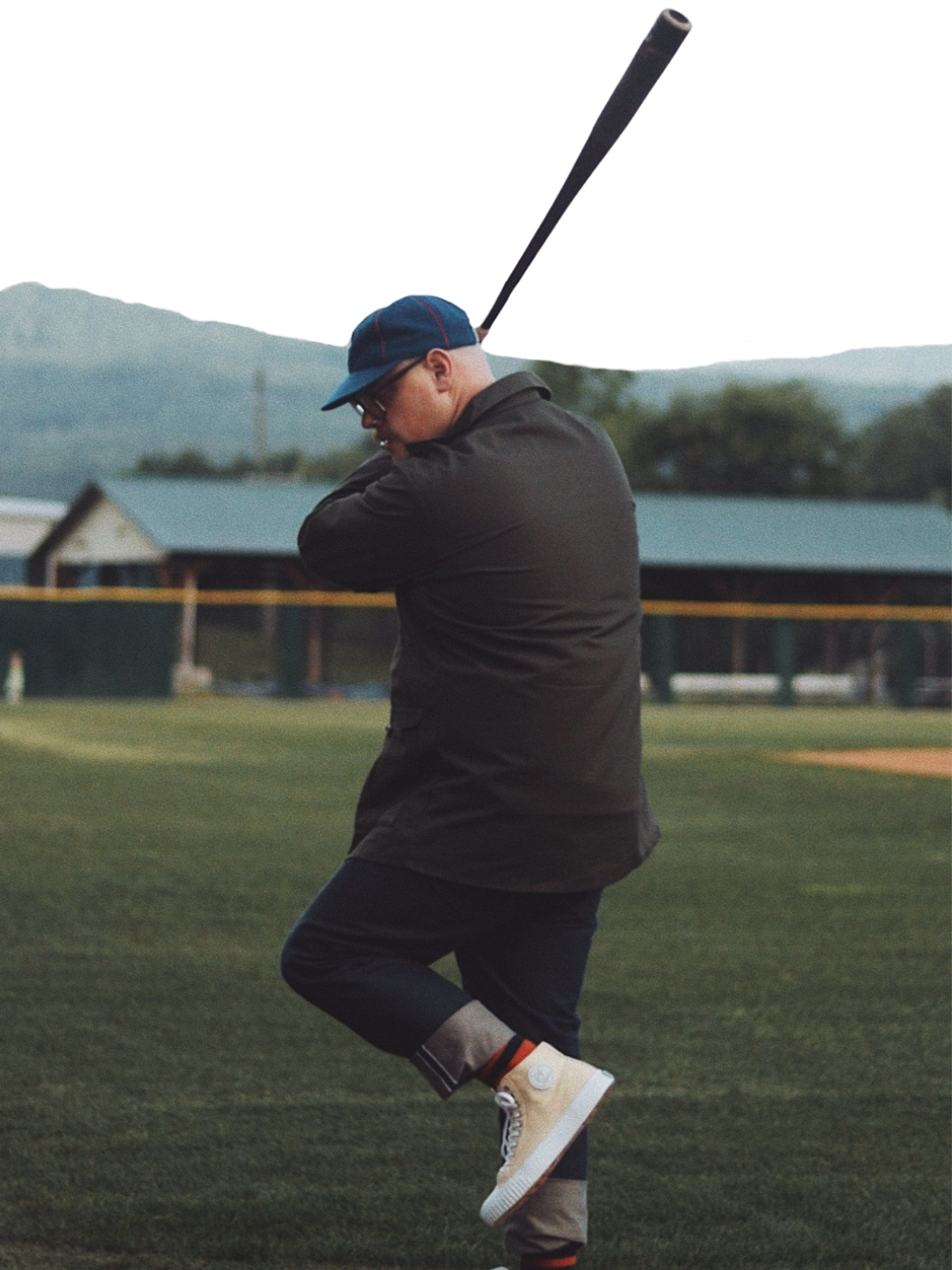 Man in a navy cap and dark jacket lifts one leg in a batting stance with a bat raised on a grassy baseball field with mountains behind.
