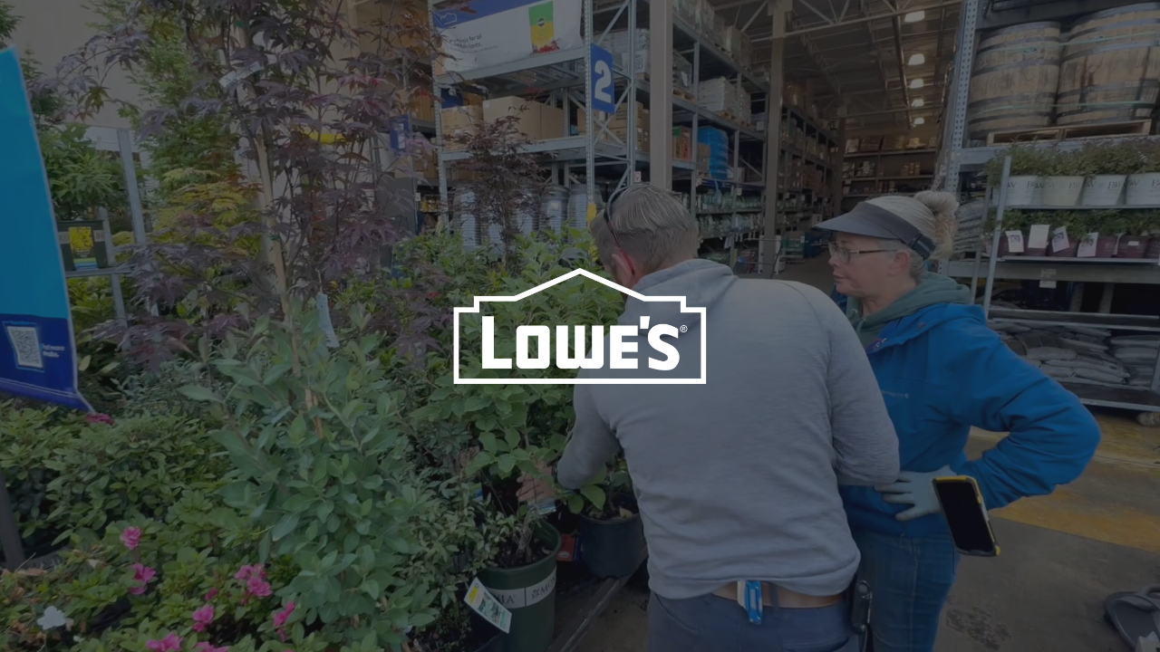 Two shoppers browse plants at a Lowe's home improvement store garden center with the Lowe's logo displayed.