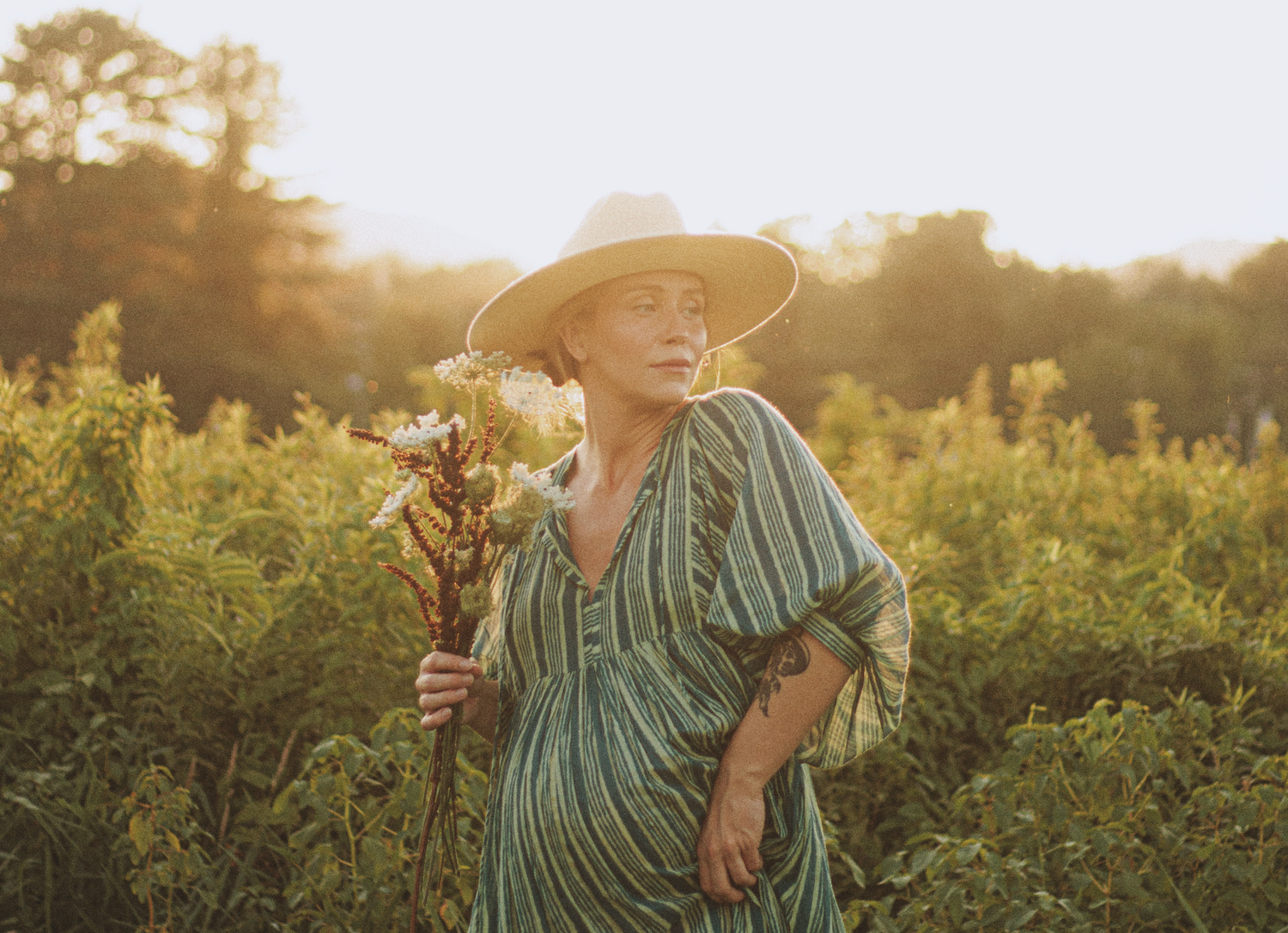 Woman in straw hat and green striped dress holding wildflowers in a sunlit garden field at golden hour.