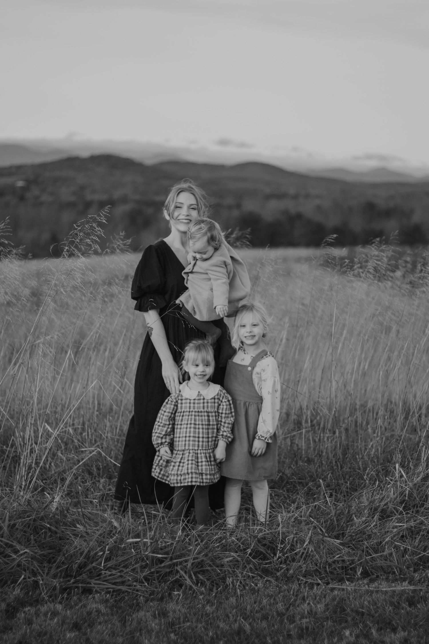 Black and white photo of a mother holding a baby with two young daughters standing in a field with mountains behind.