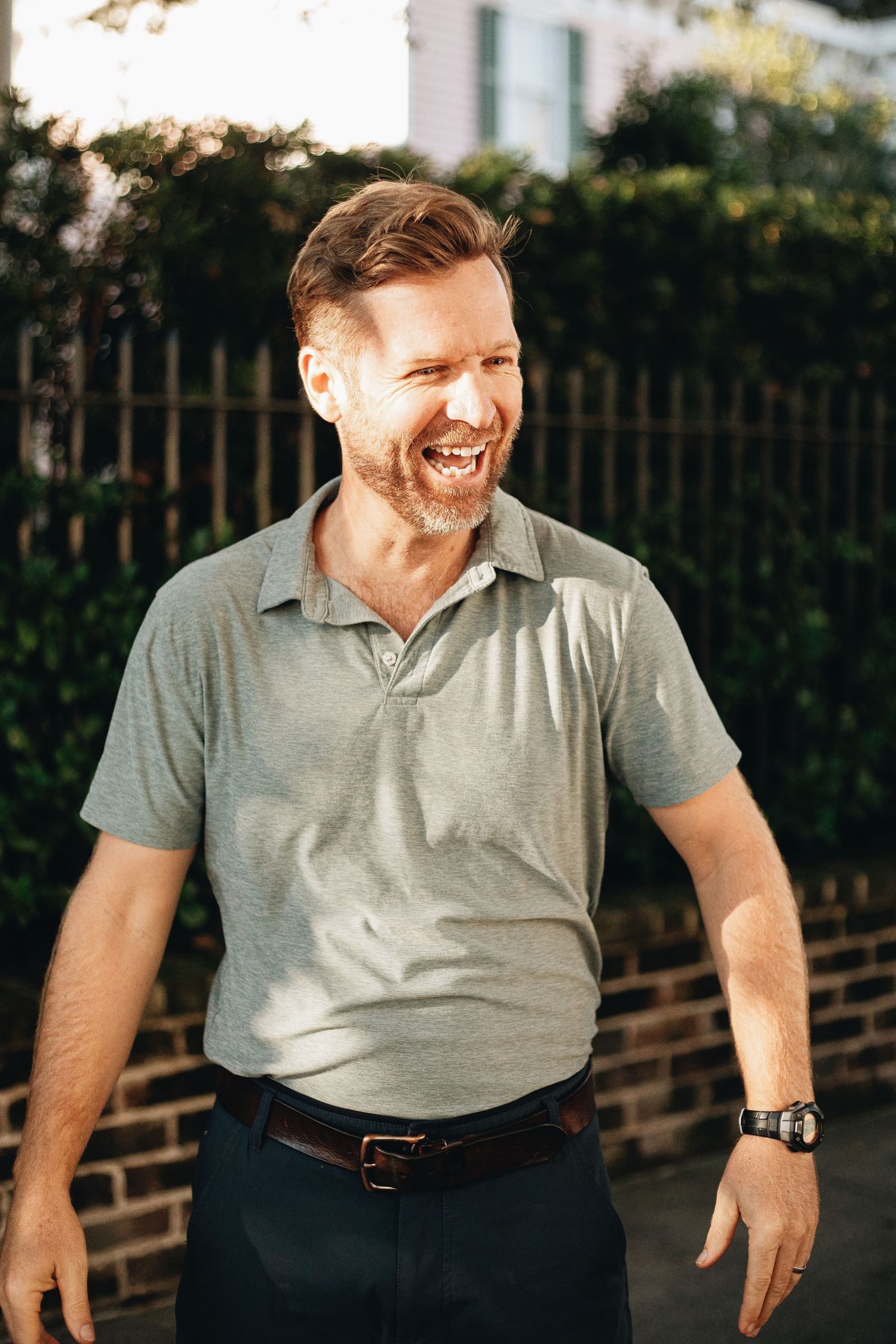 A laughing middle-aged man in a gray polo shirt and dark pants stands outdoors near a brick wall and green hedges.