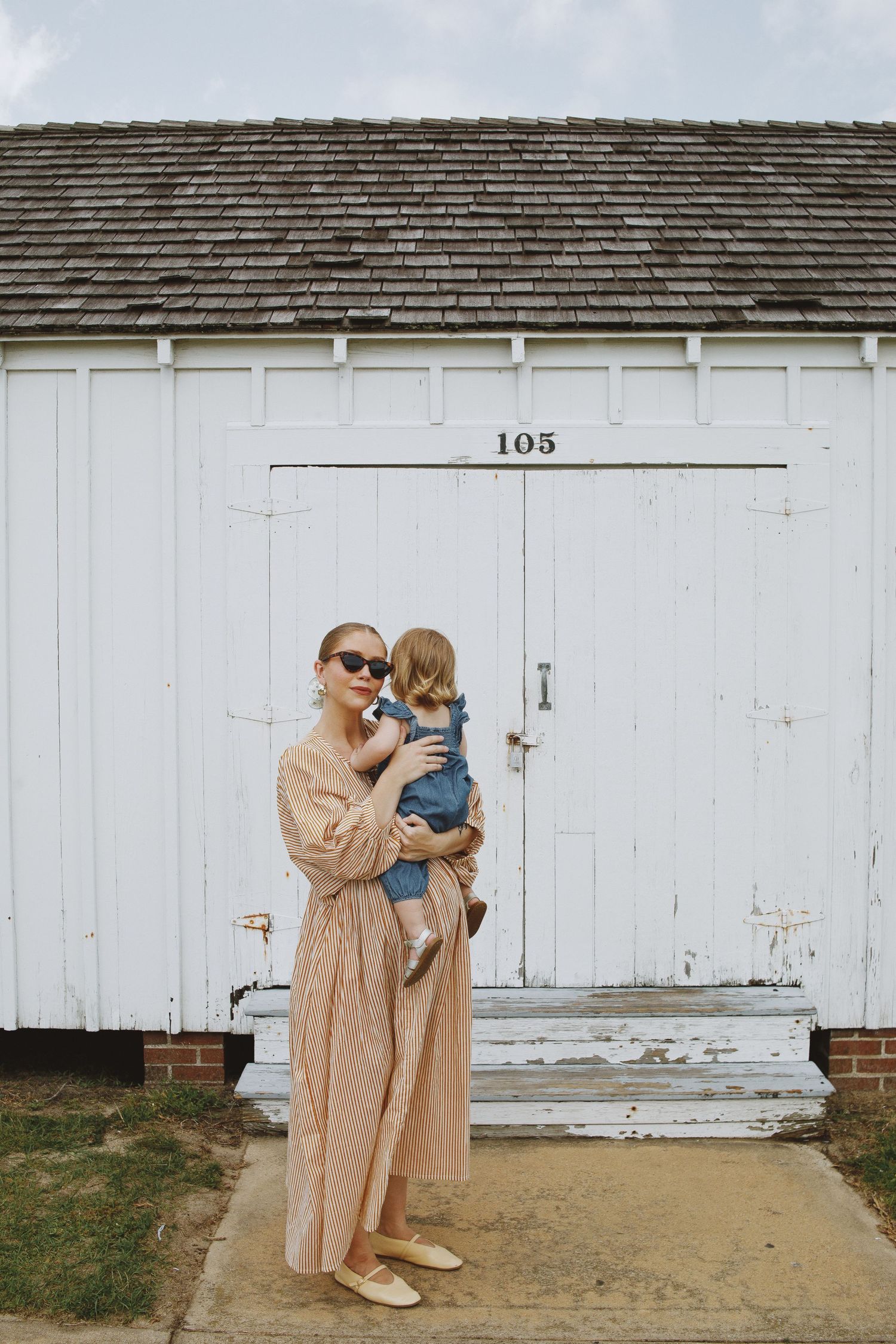 Woman in beige maxi dress holding toddler in denim outfit, standing before white barn door numbered 105.