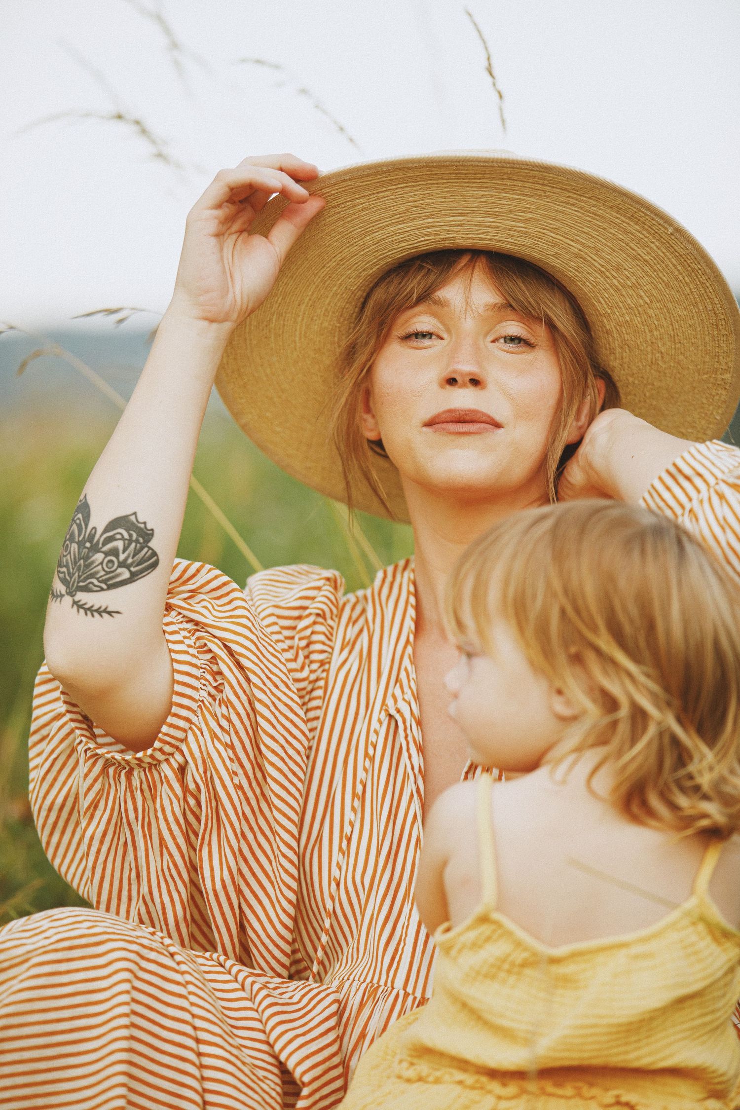 A woman in a straw hat and striped dress with a butterfly tattoo sits outdoors with a young blonde child in a yellow dress.