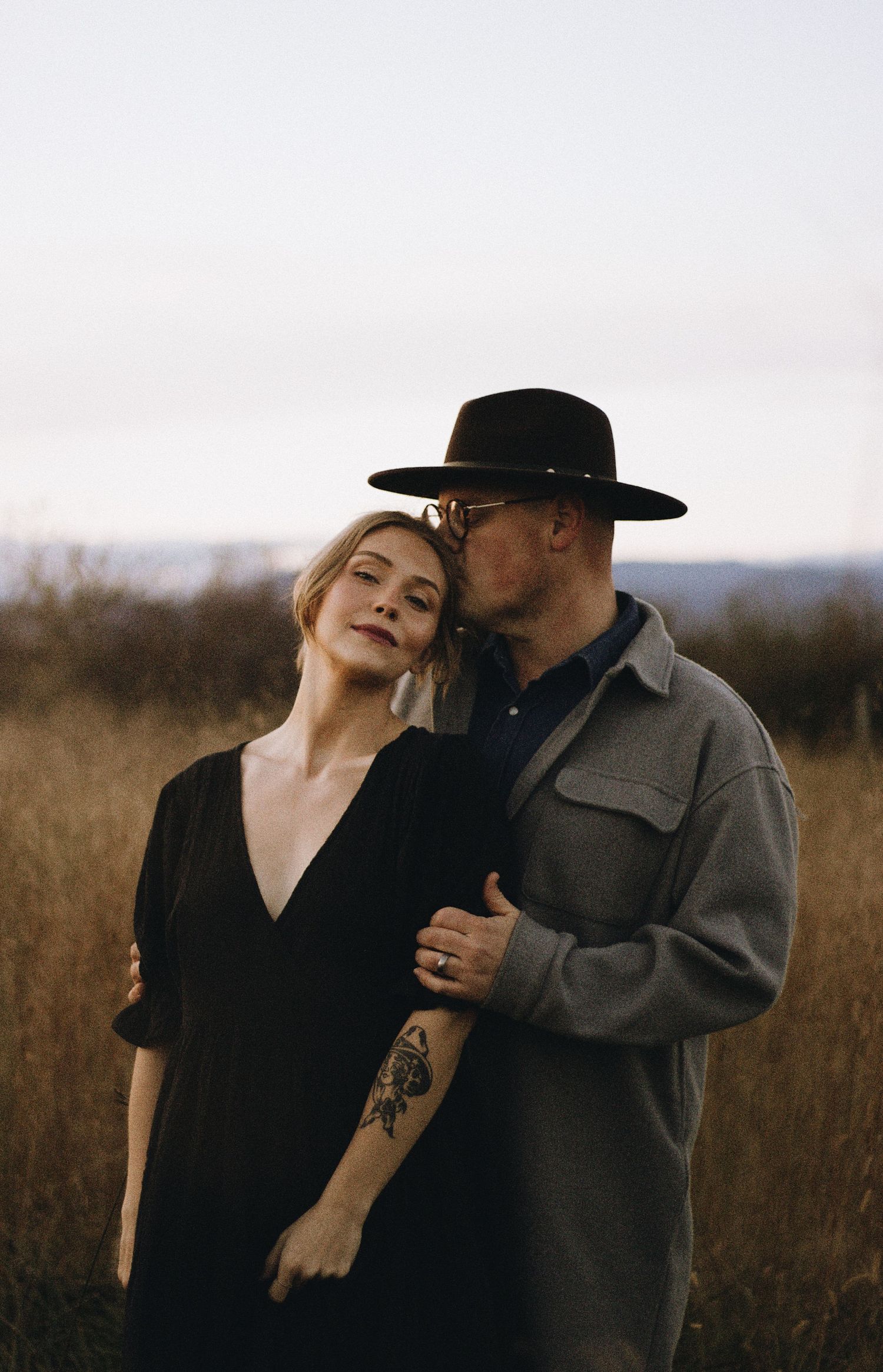 Couple shares intimate moment in field at dusk, with woman in black dress and man wearing wide-brimmed hat.