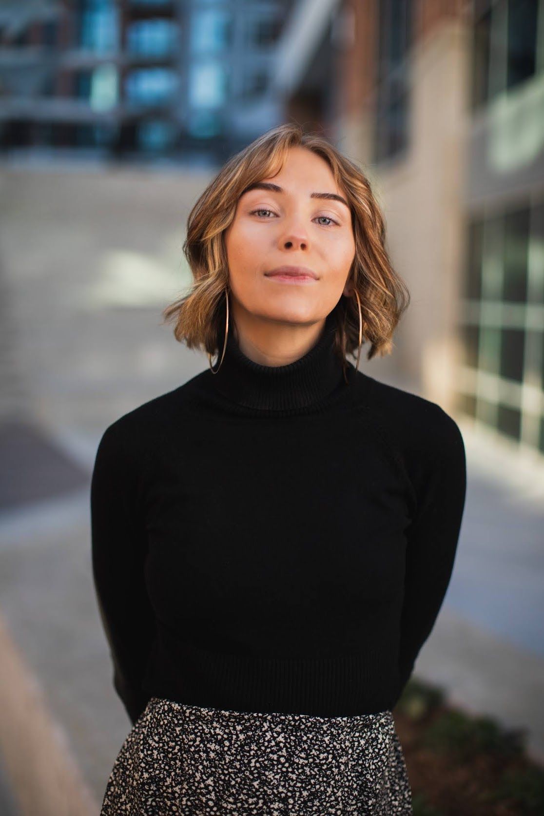 Young woman with short blonde hair wearing a black turtleneck sweater and floral skirt standing outdoors.