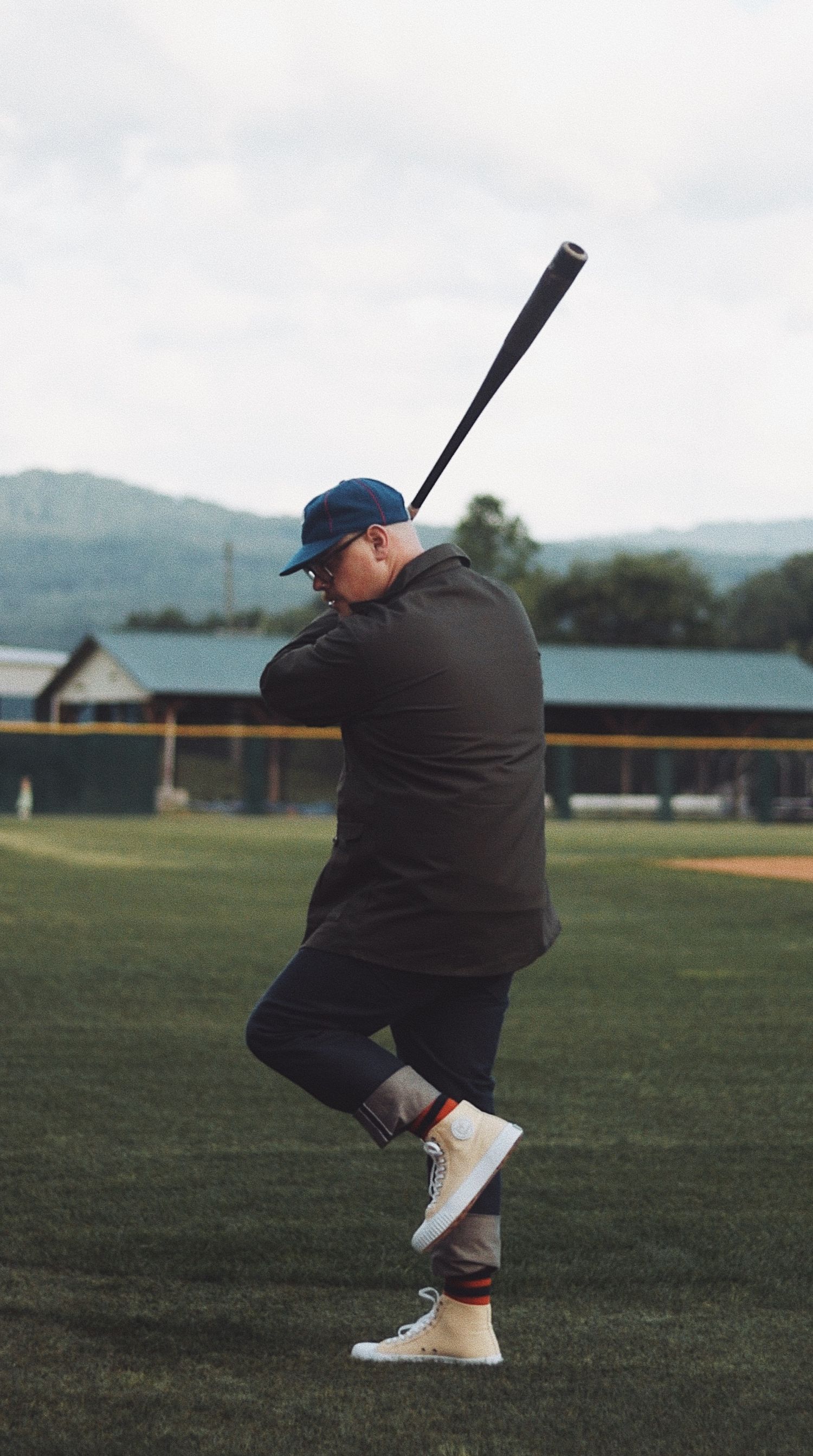 Man in dark jacket and blue cap swings a baseball bat on a green field with mountains in the background.