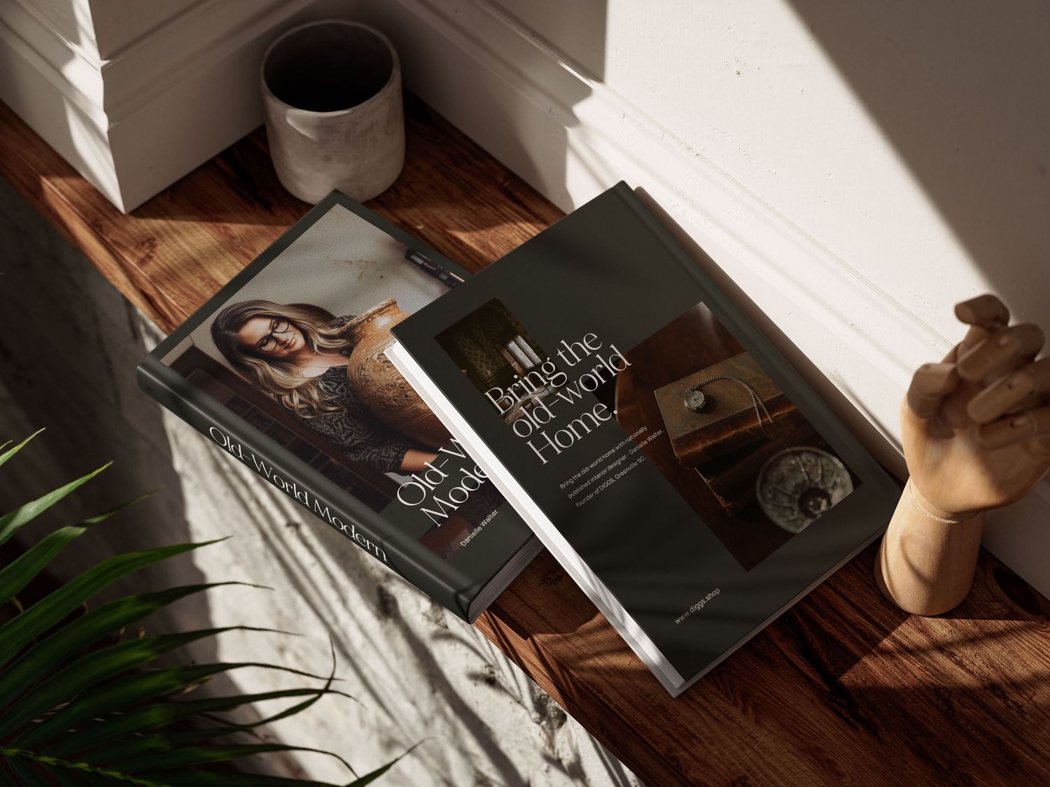 Two coffee table books on hardwood floor with sunlight, a ceramic mug, and a wooden hand figurine nearby.