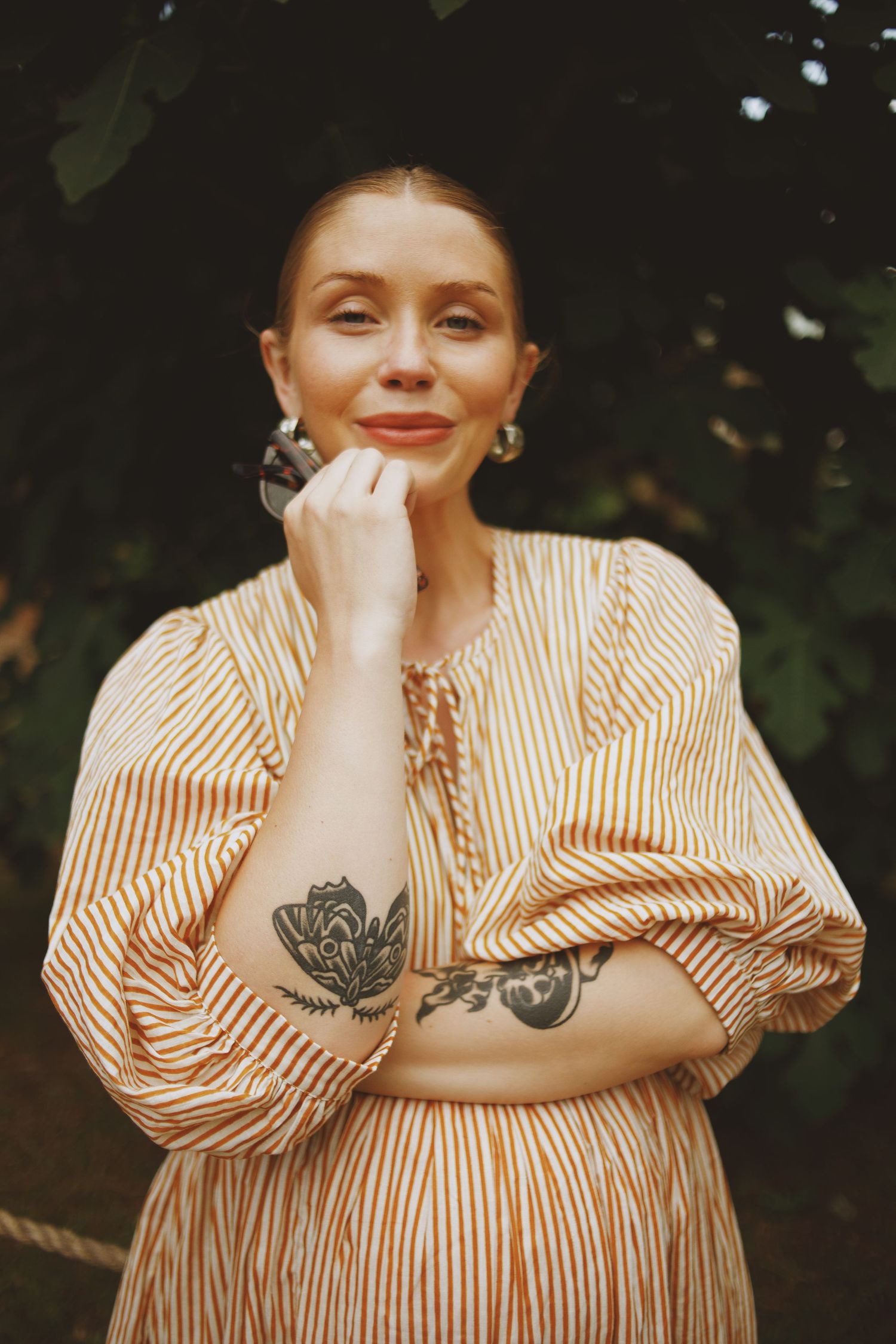 Woman in striped orange dress with floral tattoos smiling outdoors among green foliage.