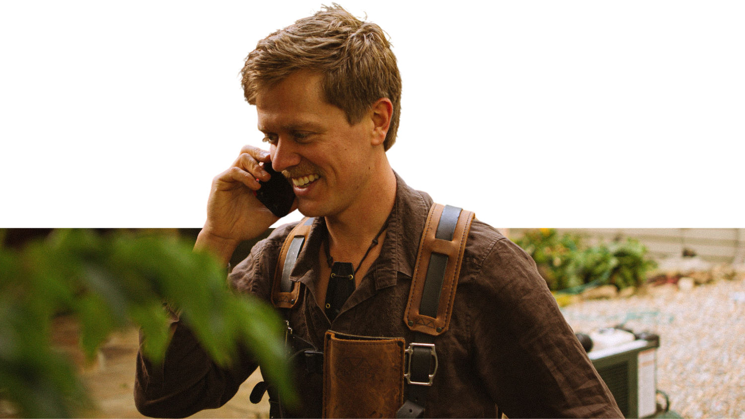 Smiling man in a brown work shirt and leather tool suspenders talks on a cell phone while standing outdoors near green plants.