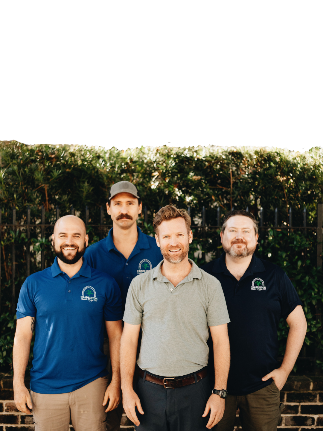 Four smiling men in matching polo shirts with a company logo pose together outdoors in front of a green hedge.