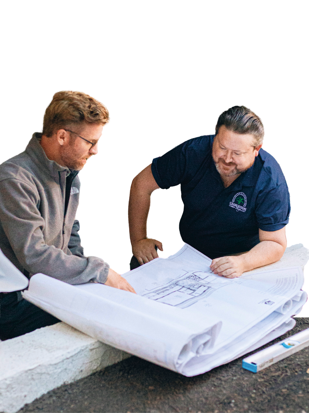 Two men in casual work attire review rolled architectural blueprints together while sitting outdoors on a concrete ledge.
