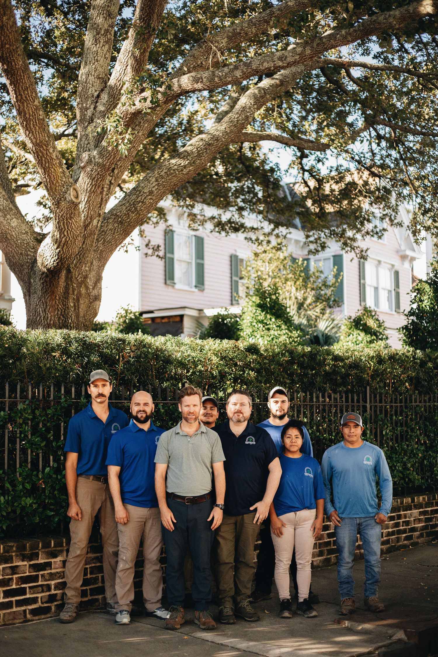 A team of eight landscaping professionals in blue polo shirts posing outdoors beneath a large oak tree.
