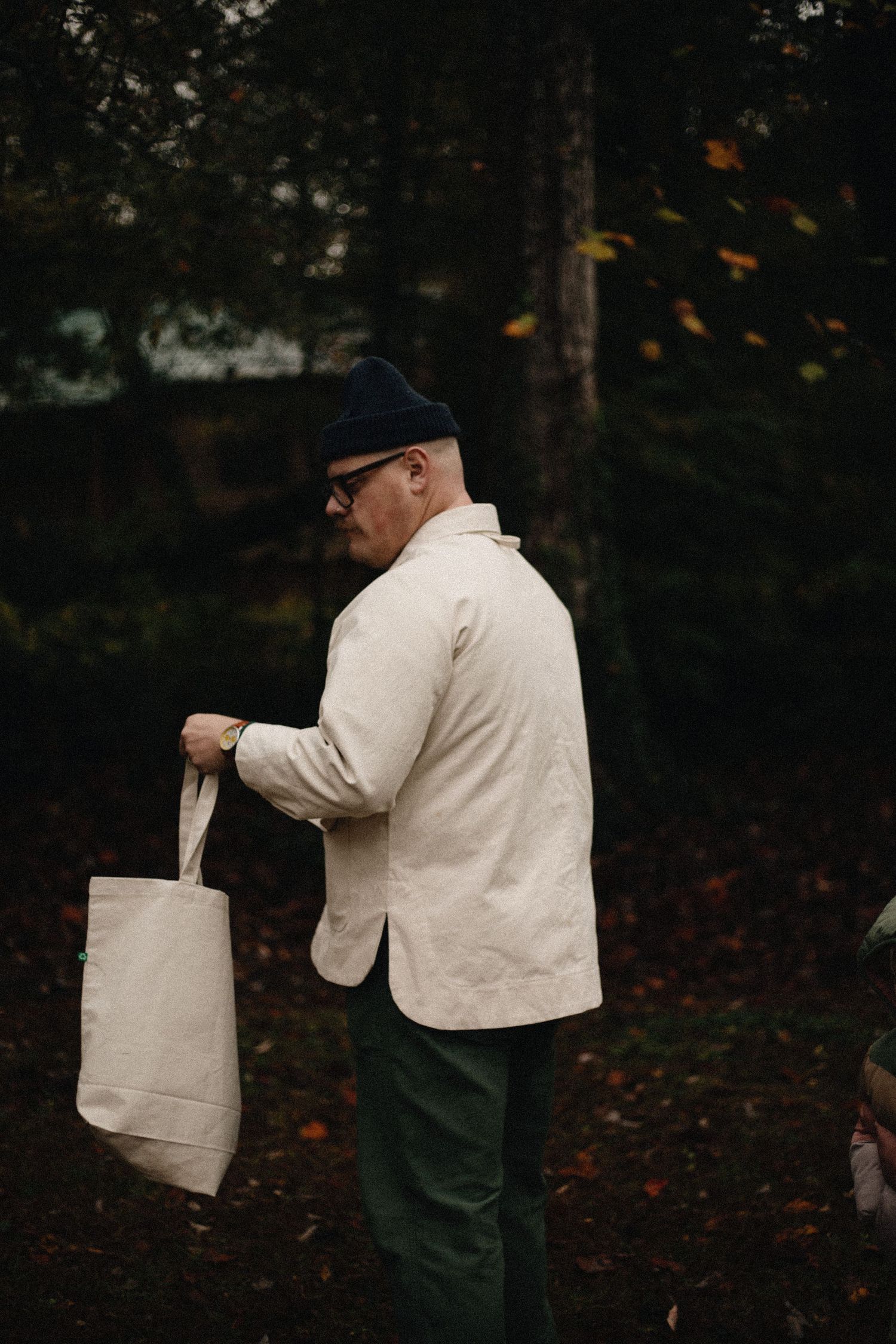 Man in a dark beanie, glasses, cream jacket and green pants holds a canvas tote bag while standing in an autumn forest with fallen leaves.