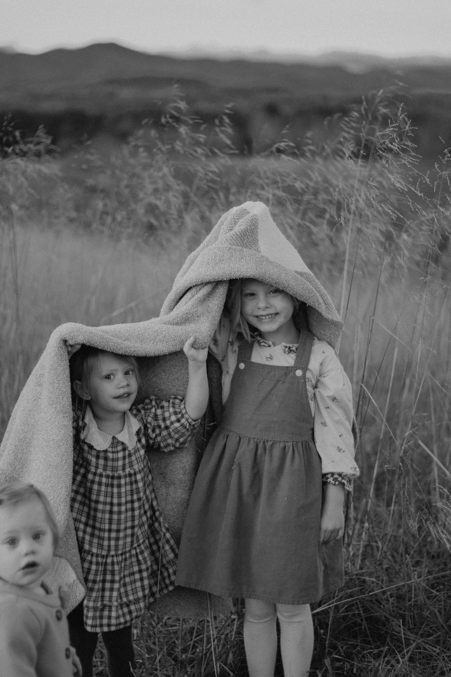 Three young girls playing outdoors in a field, one wearing a hooded blanket, captured in black and white.
