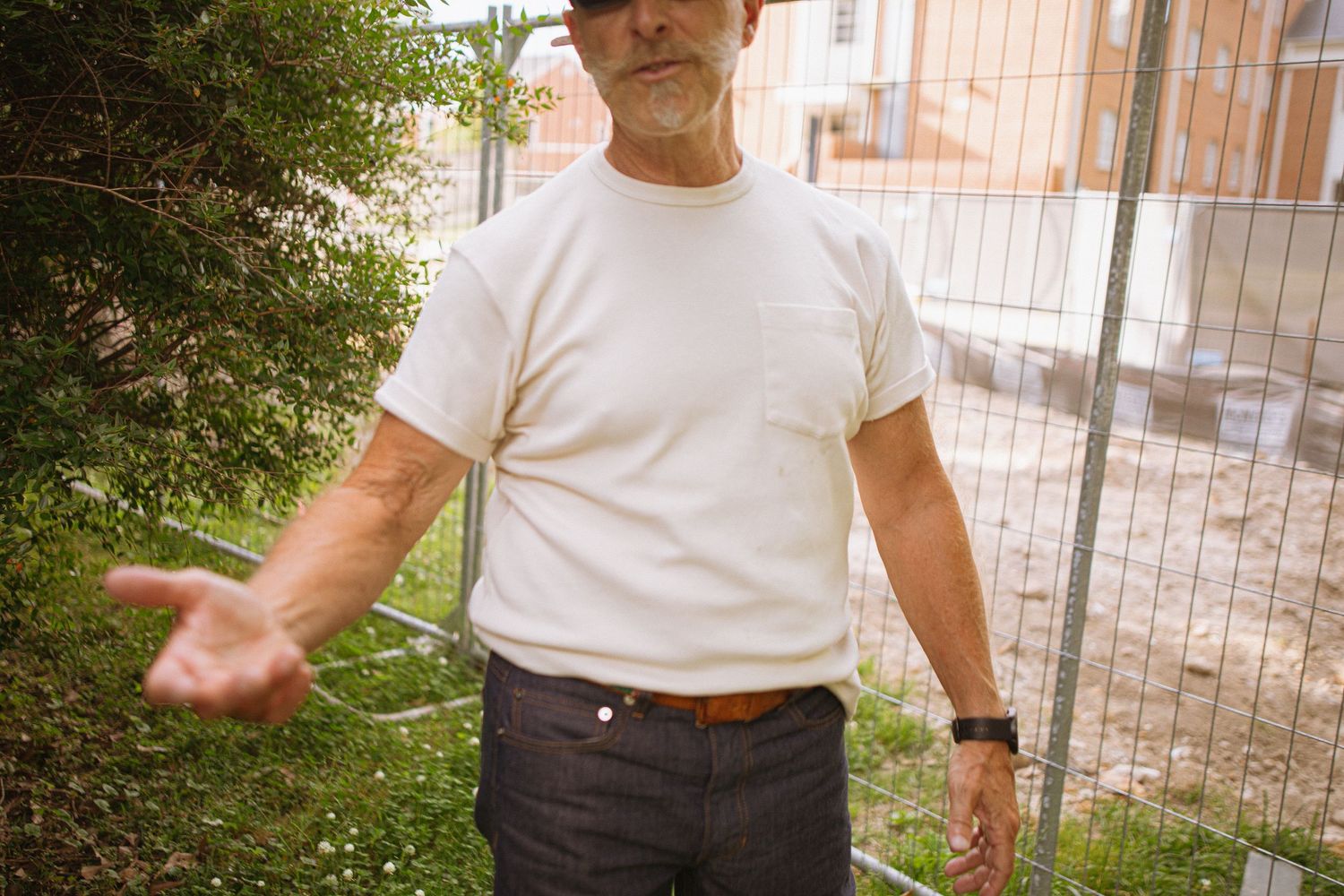 An older man in a white t-shirt and jeans gestures with his hand near a construction fence outdoors.