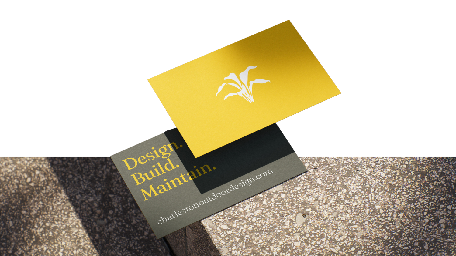 Two business cards, one yellow with a leaf logo floating above a dark card reading Design. Build. Maintain., on a textured concrete surface.