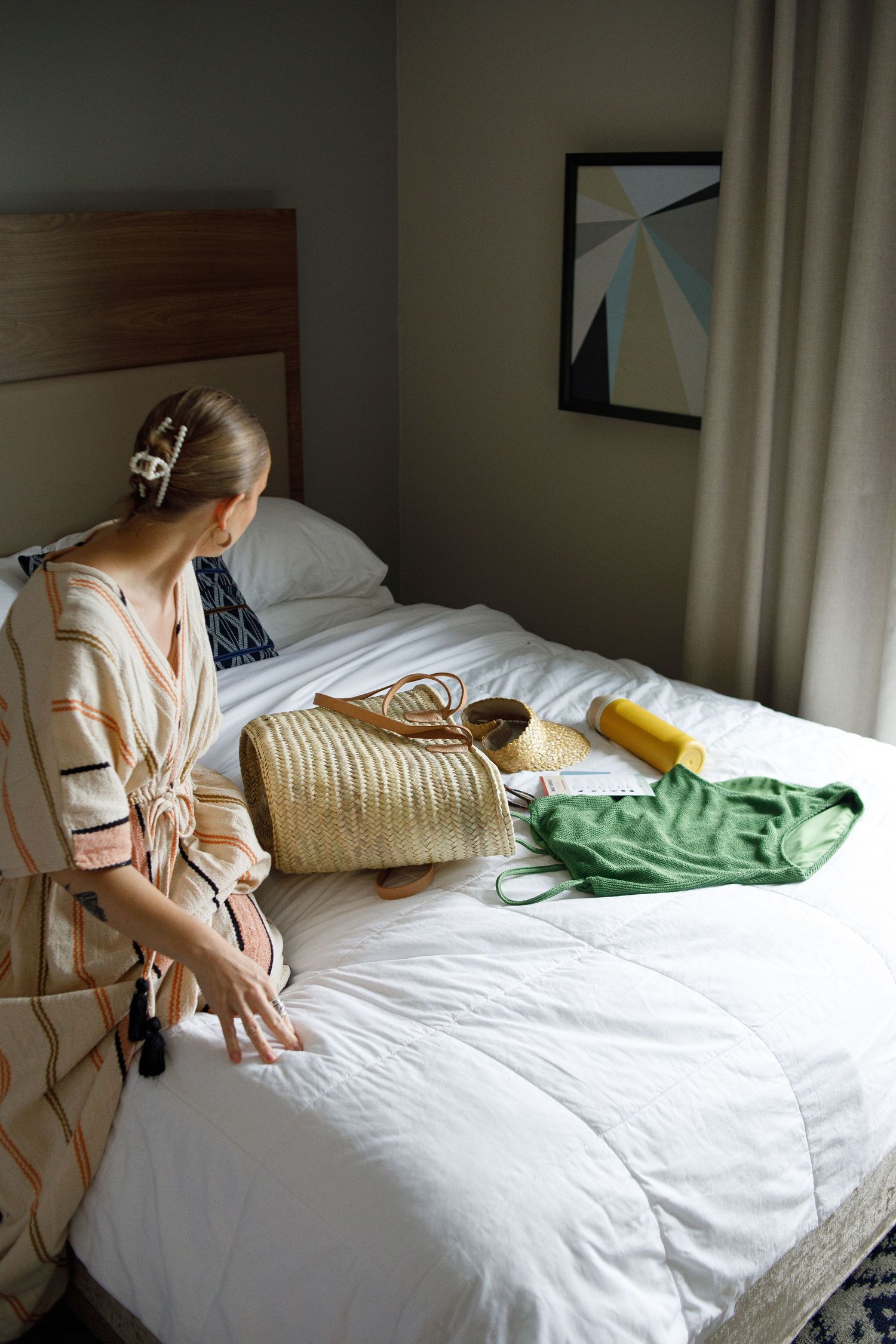 Woman in floral robe sitting on white hotel bed with straw bag, green swimsuit, and yellow item laid out.