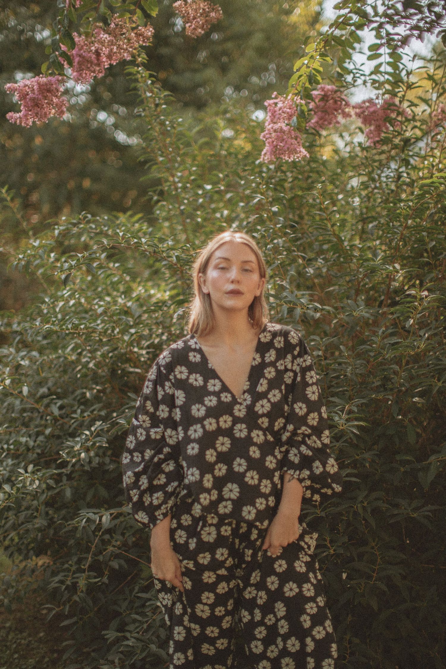 Woman in floral black dress standing among lush green shrubs with pink blooms in soft natural light.
