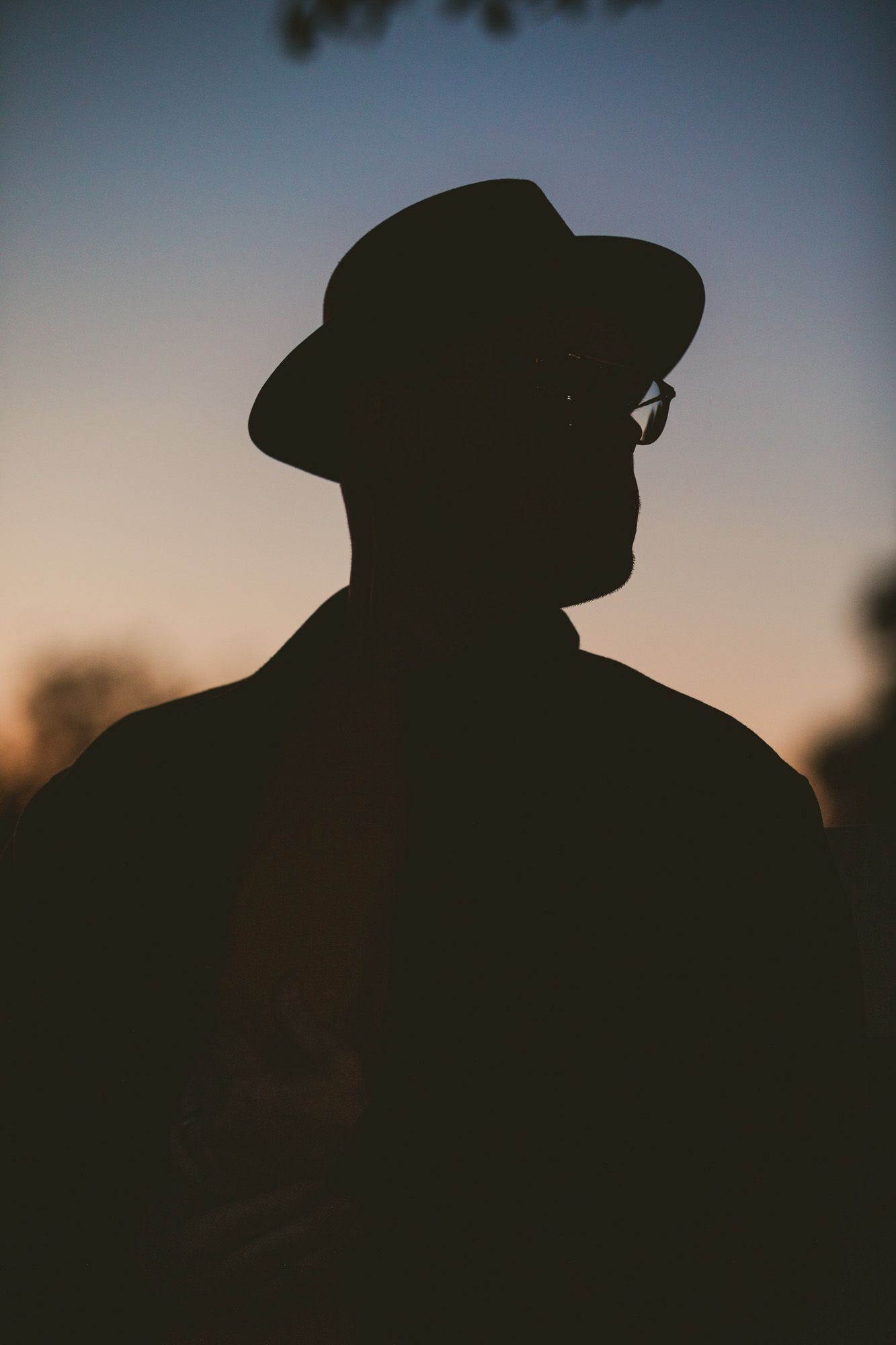 Silhouette of a man wearing a wide-brim hat and glasses against a warm sunset sky.