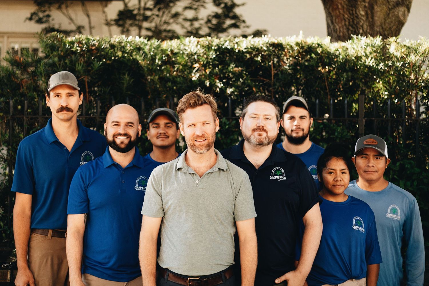 A team of eight men and women in matching blue and navy polo shirts and caps pose together outdoors in front of green hedges.