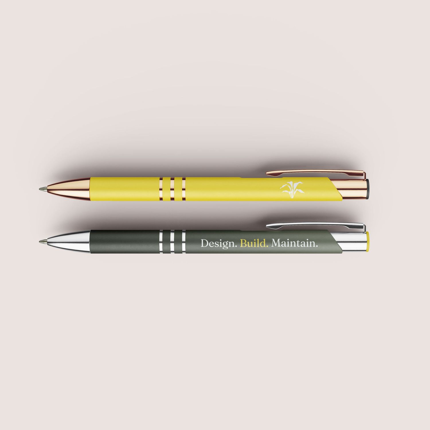 Two metal ballpoint pens in yellow and olive green with rose gold and silver accents, engraved with logos and text.