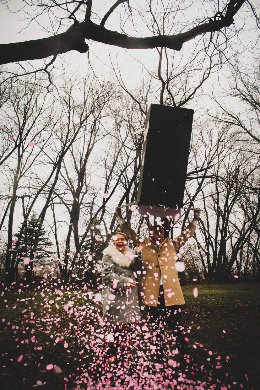 Two people joyfully toss pink confetti in a park with bare trees during a gender reveal celebration.