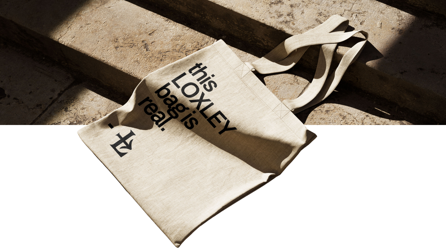 Natural canvas tote bag with black text reading this LOXLEY bag is real, resting on sunlit stone steps with soft shadows.