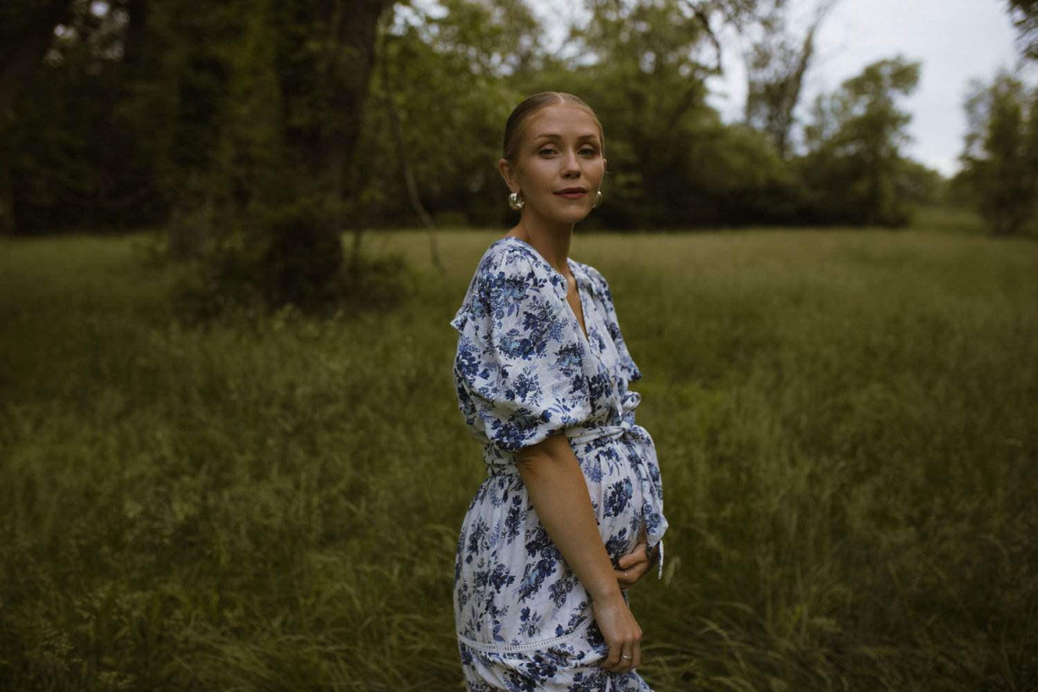 Pregnant woman in blue floral dress standing in lush green field, posing for maternity photo session outdoors.