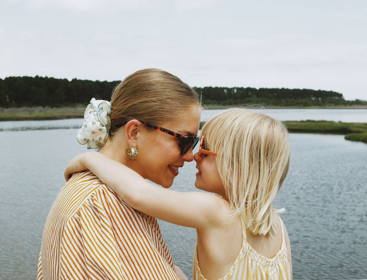 A mother and young blonde daughter touch foreheads affectionately outdoors near a serene lake with lush greenery behind them.
