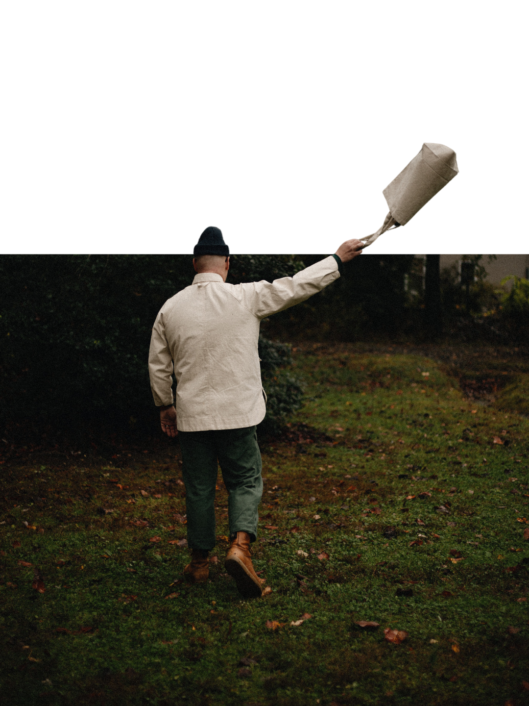 Man in a cream jacket, beanie and brown boots stands on a leafy lawn, raising a tan tote bag high in one outstretched arm.