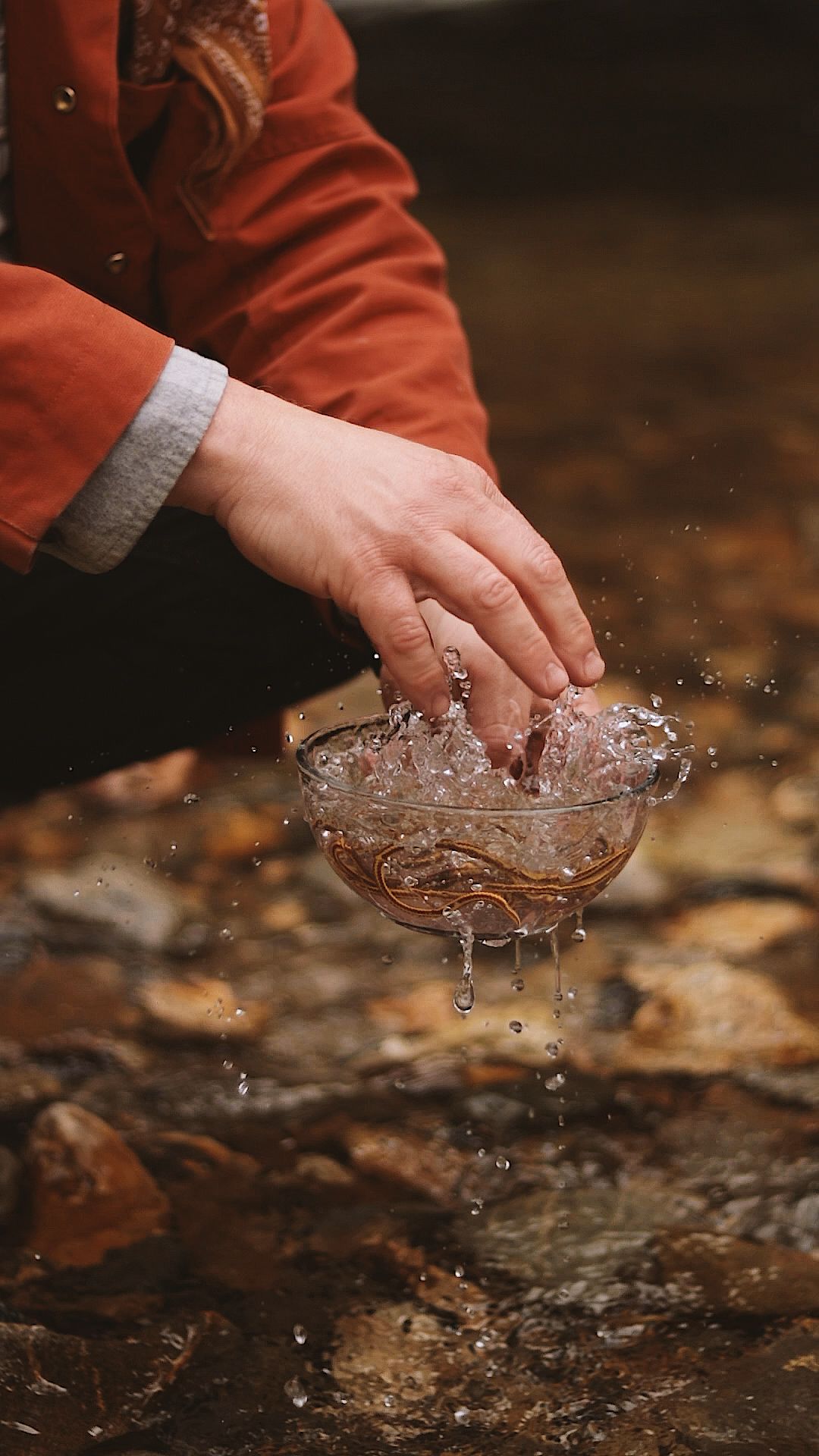 Hands scooping fresh water from a rocky stream, with splashing droplets, wearing a red jacket outdoors.