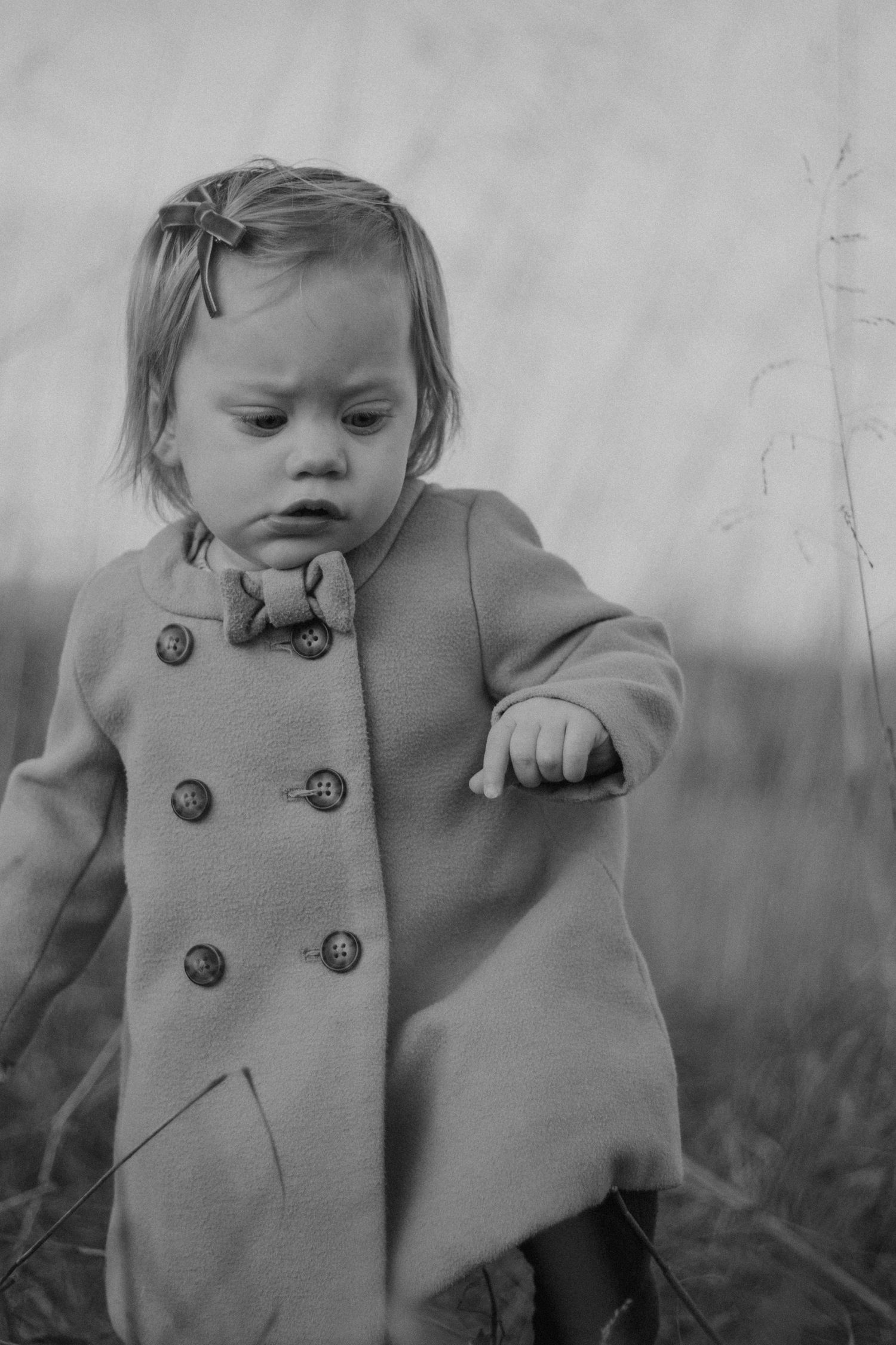 Black and white photo of a toddler girl in a double-breasted coat with bow hair clip standing in a field.