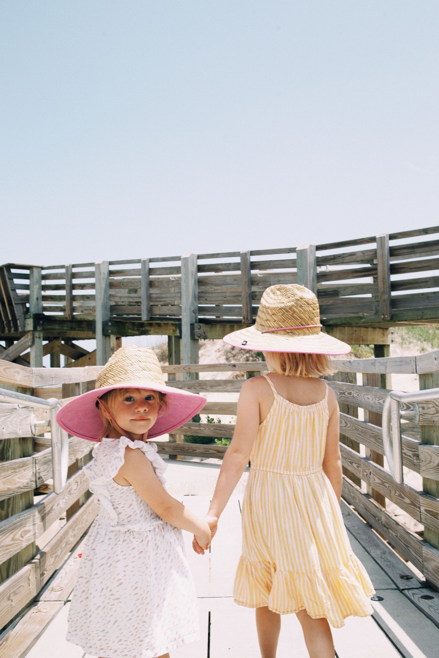Two young girls in straw hats and summer dresses hold hands on a sunny wooden boardwalk.