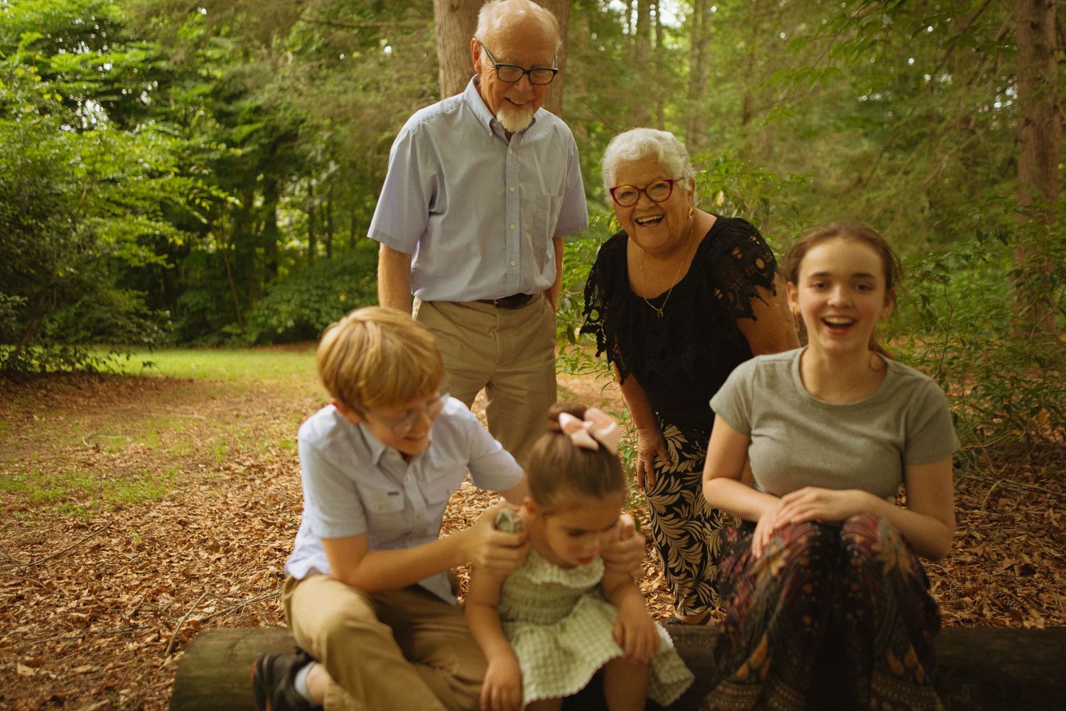 Family of five poses outdoors in a wooded area, with grandparents standing behind three young children sitting on a log.