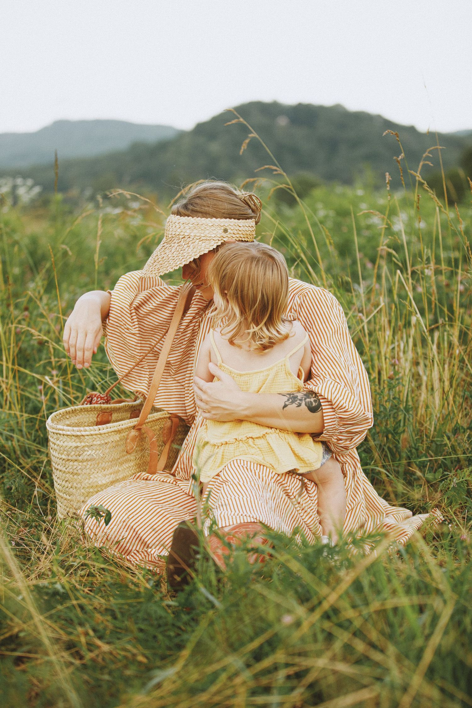 Mother and child sitting in tall grass field with wicker basket, mountains in background, wearing striped and yellow dresses.
