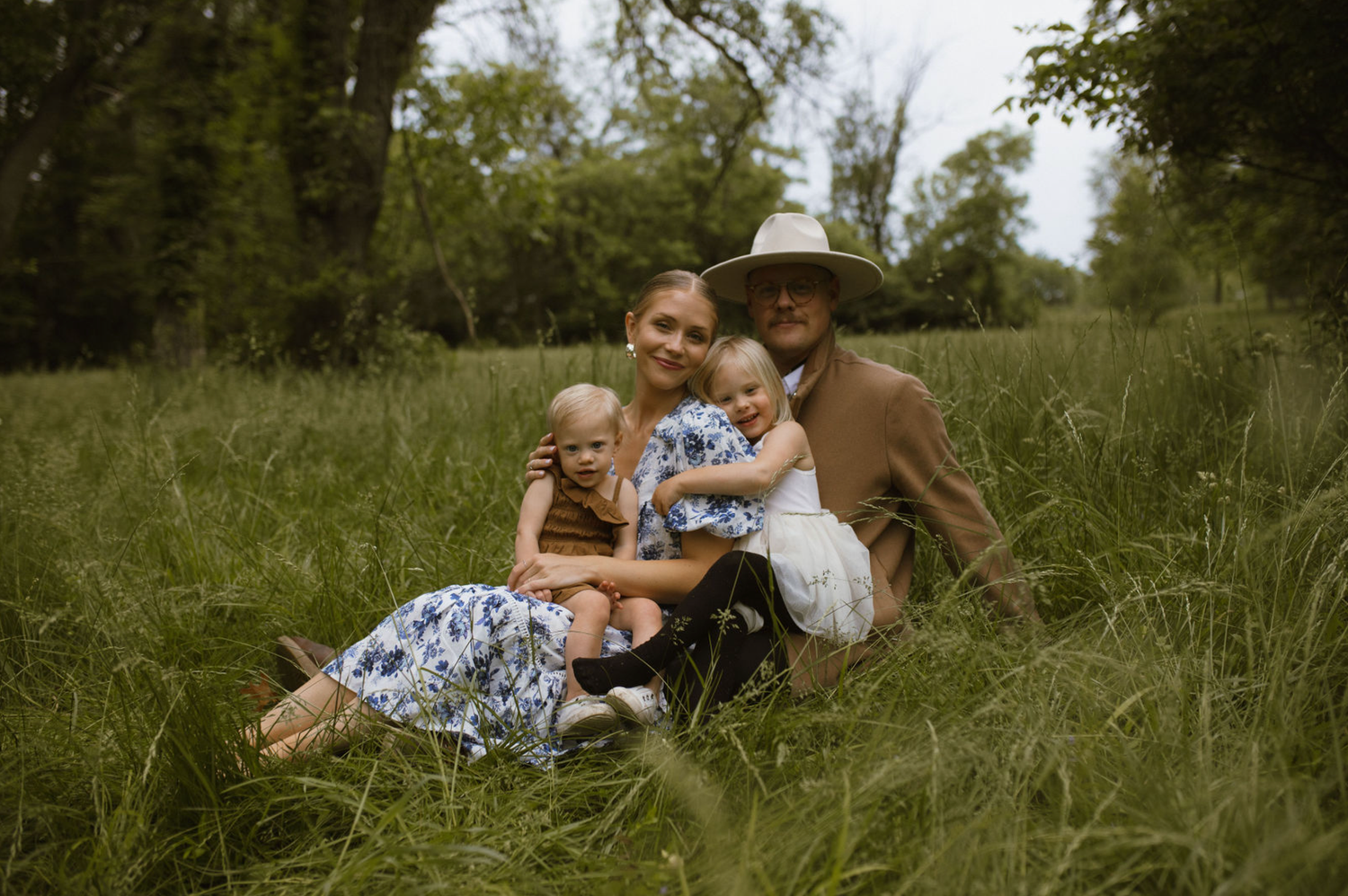 Family of four sitting in lush green meadow, parents with two young children in floral dresses, smiling outdoors.
