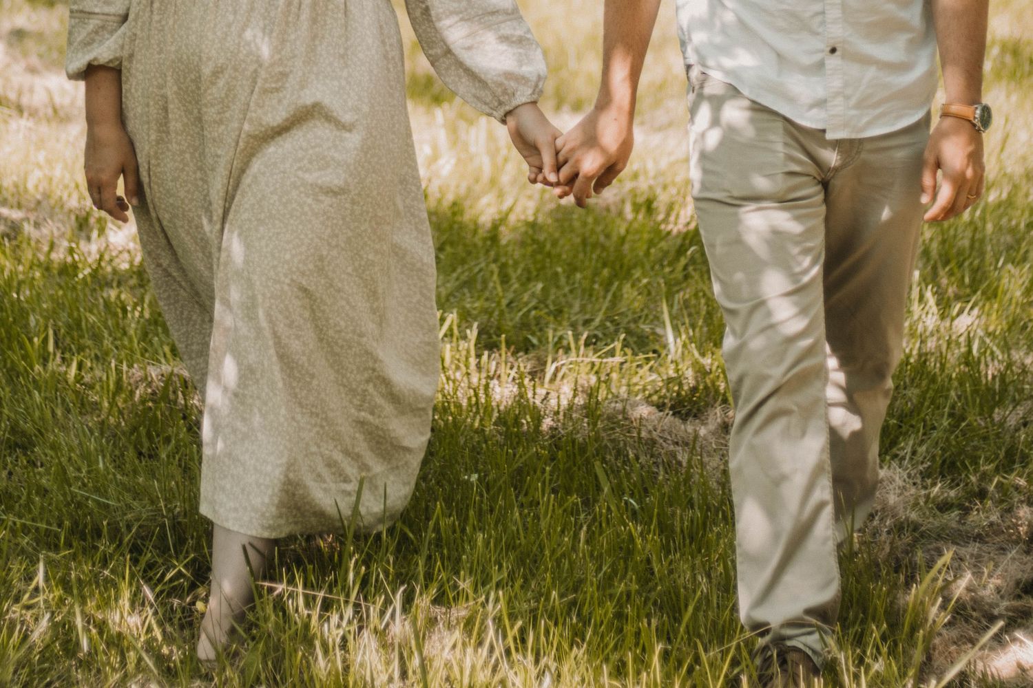 Couple holding hands walking through a sunlit green field, wearing neutral tones, romantic outdoor lifestyle photography.