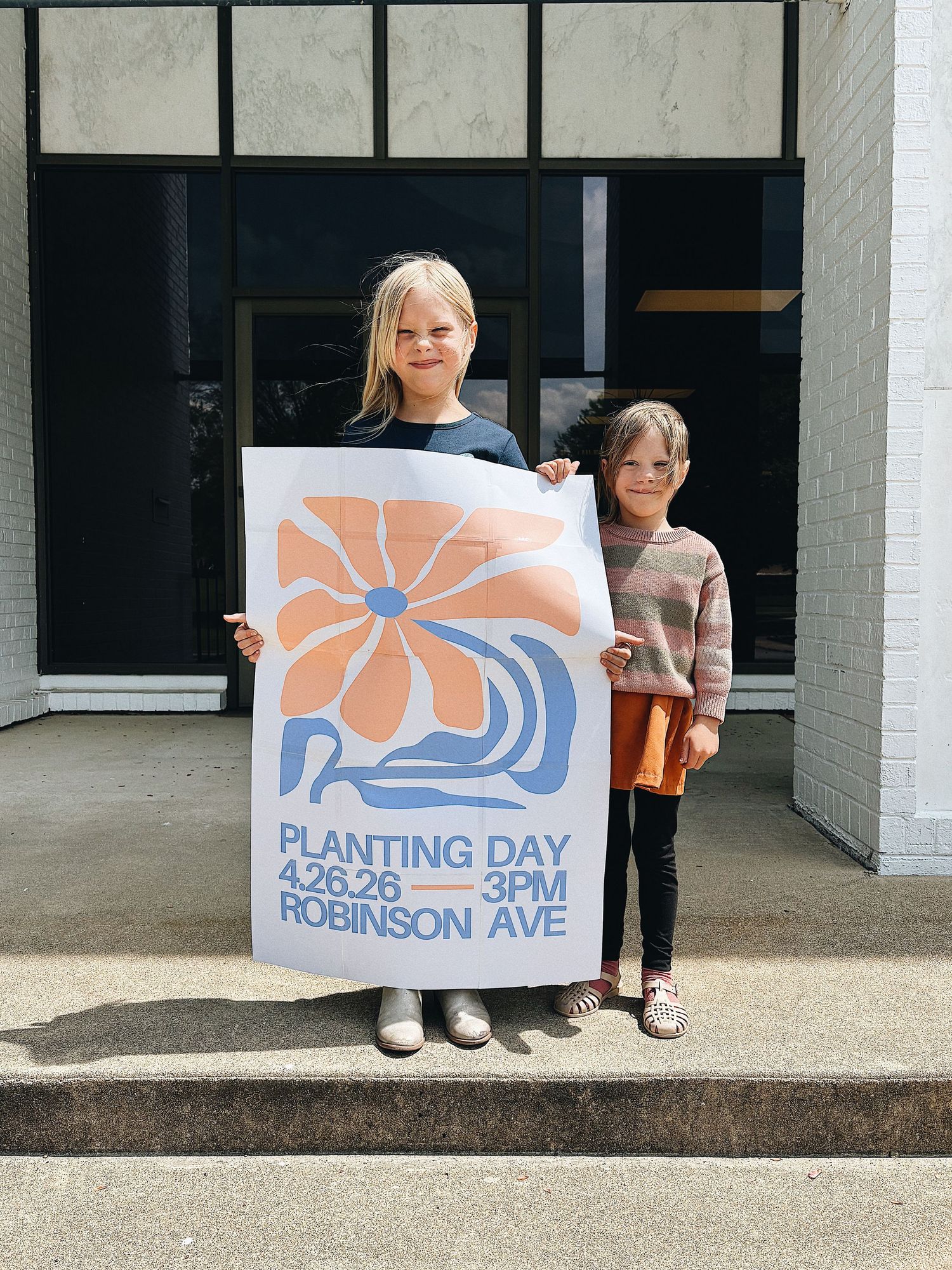 Two children holding a large poster for Planting Day event on 4.26.26 at 3PM on Robinson Ave.