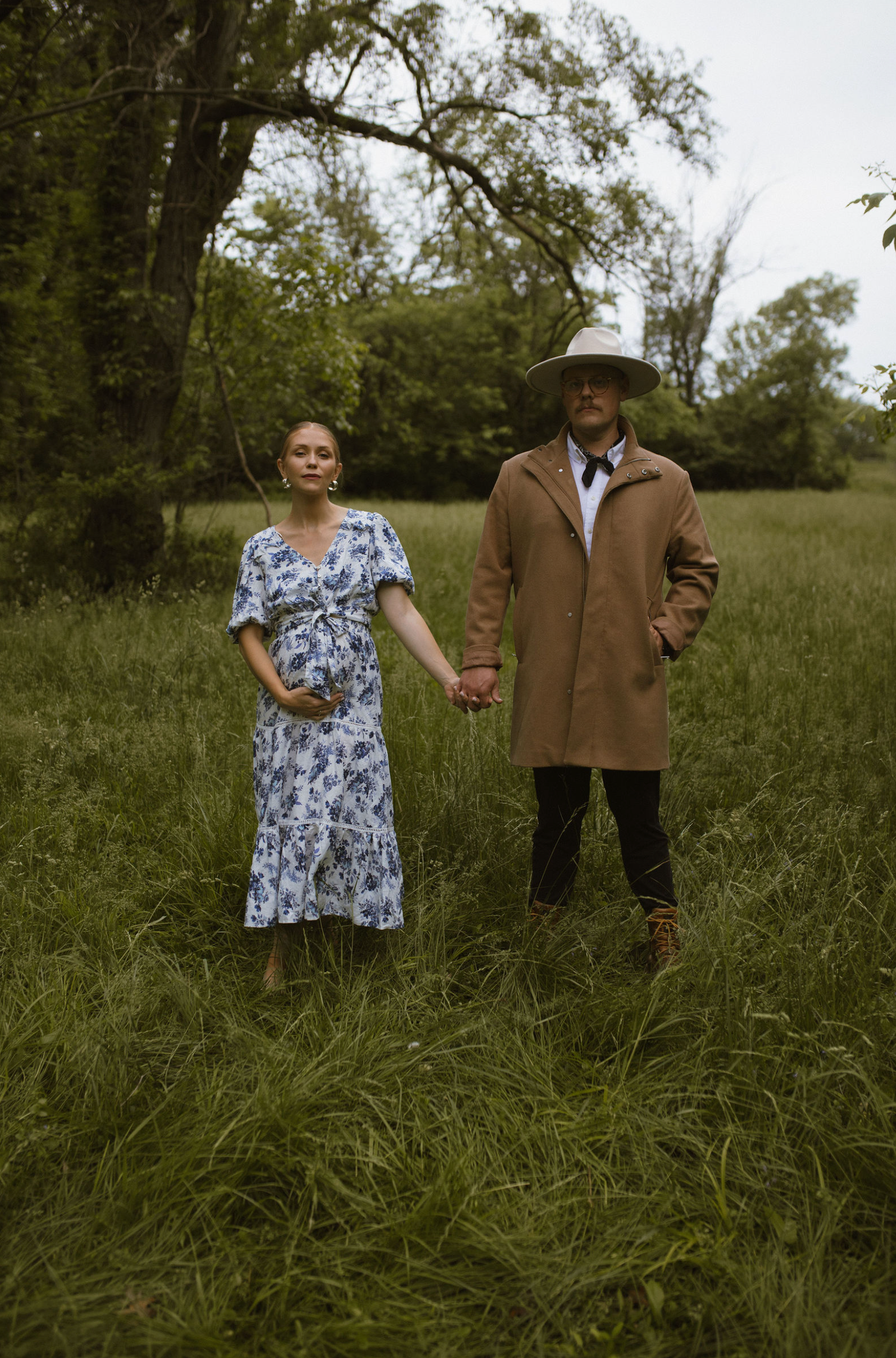 A pregnant woman in a floral dress holds hands with a man in a tan coat and wide-brim hat standing in a lush green field.