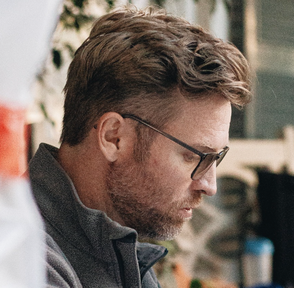 Side profile of a bearded man with swept-back hair and dark-rimmed glasses, wearing a gray fleece jacket and looking down in concentration.