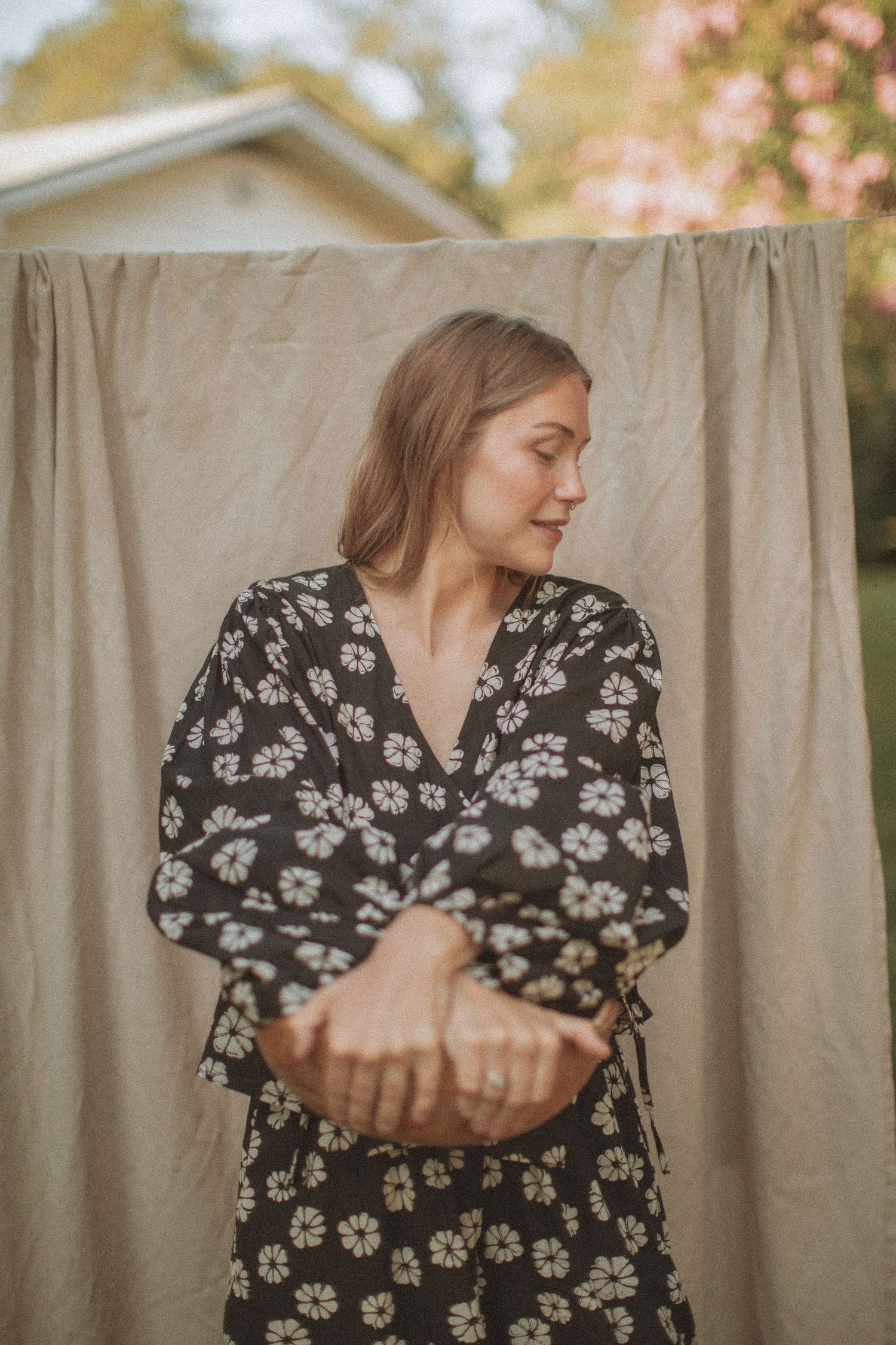 Woman in black floral wrap dress standing outdoors against a neutral linen backdrop with soft natural lighting.